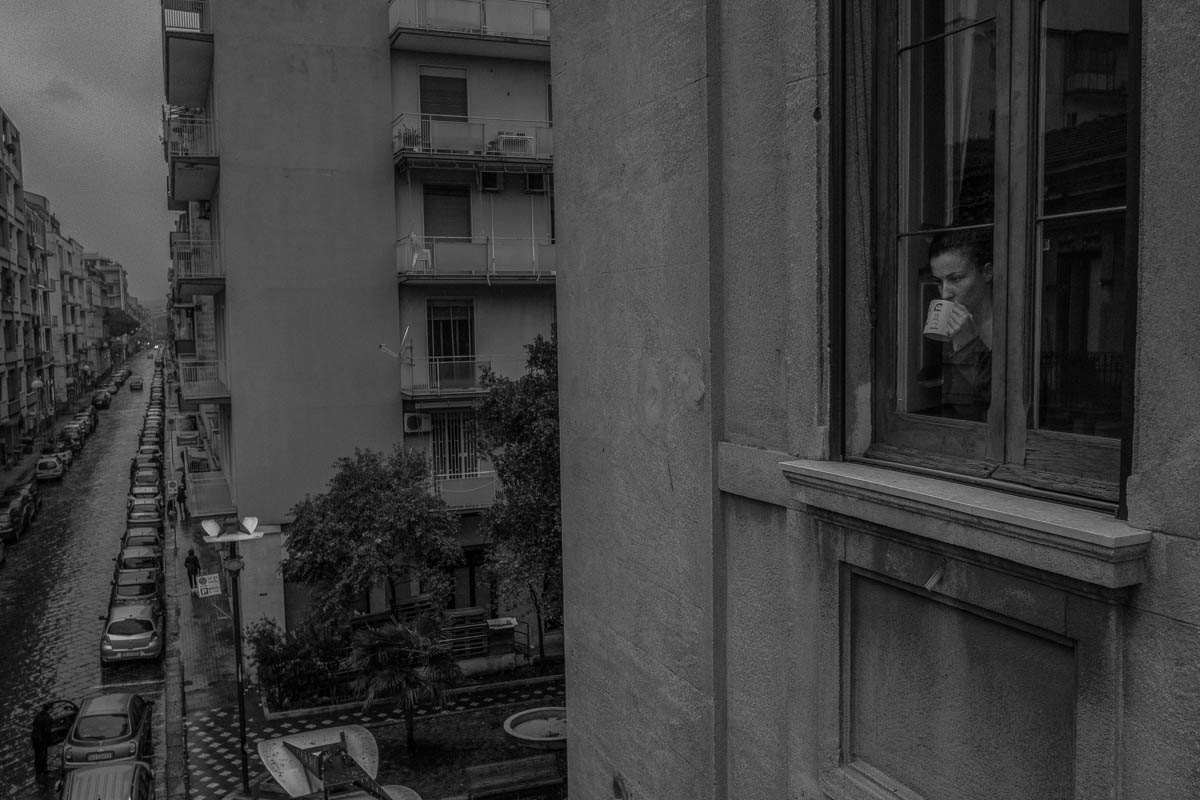 Marta at work on her laptop the window while drinking an herbal tea. In the background, some people queue in the street waiting to enter the supermarket under the house.