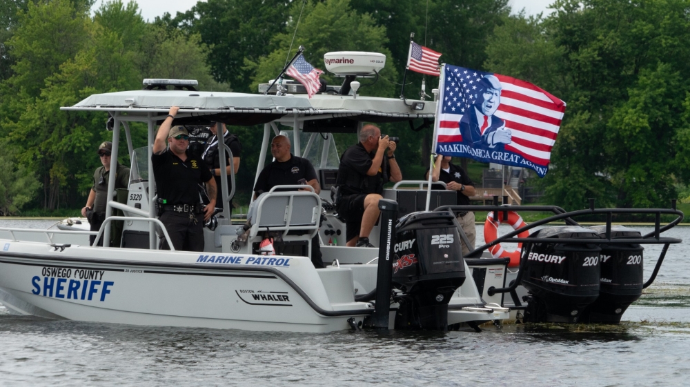 Uniformed Oswego County Sheriff’s deputies escort and cheer on a pro-Trump boat rally, flying a large ‘MAGA’ flag in Brewerton, New York