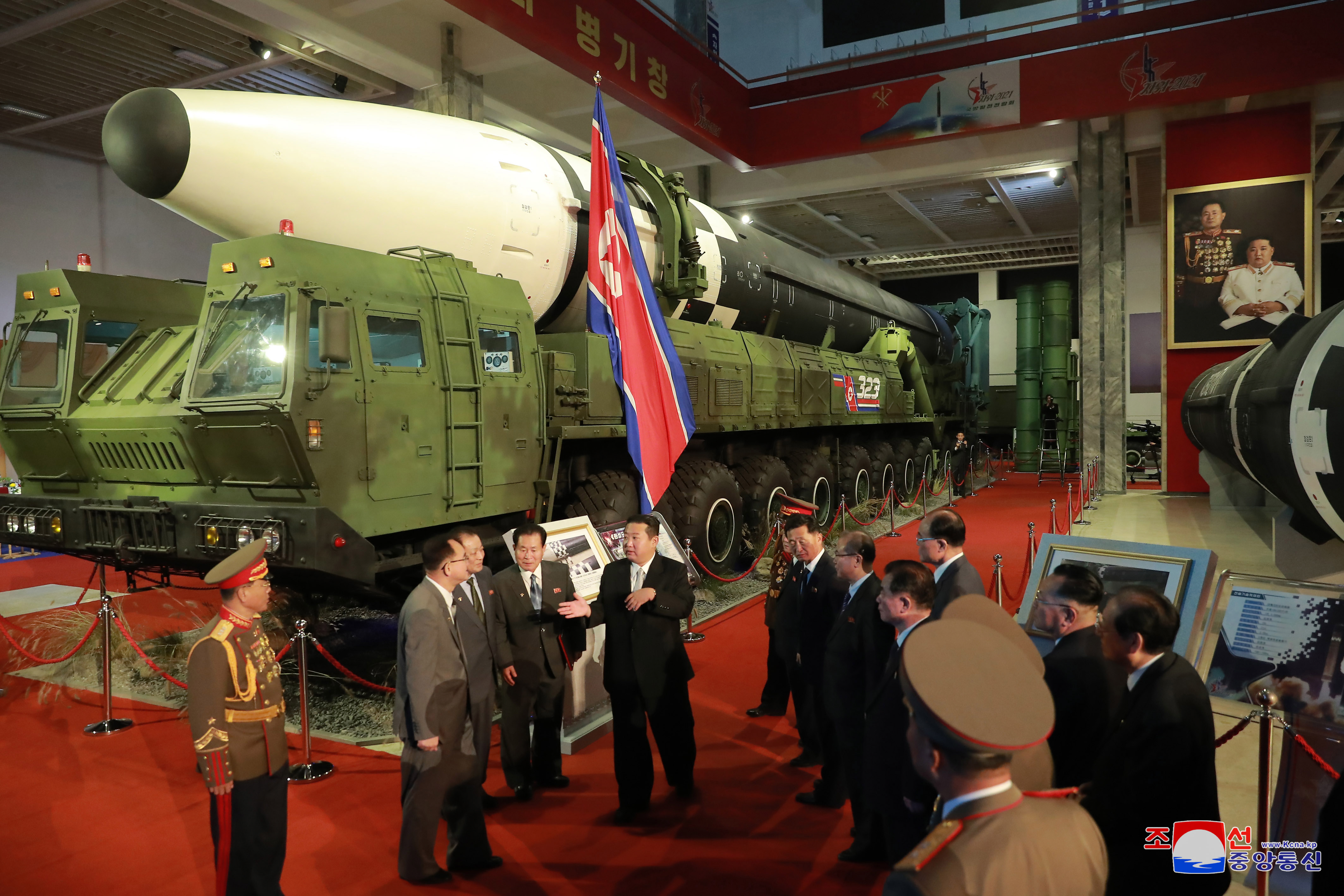 North Korean leader Kim Jong Un speaking to officials at a defence exhibition in October 2020 in front of a rocker with a white nose cone on a launcher, and a North Korean flag 