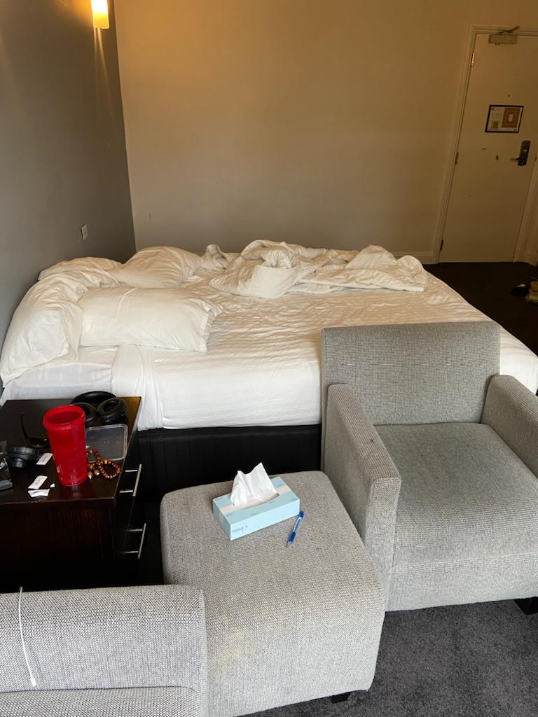 The inside of Hossein's room with an unmade ned, grey armchair and stool and beige walls