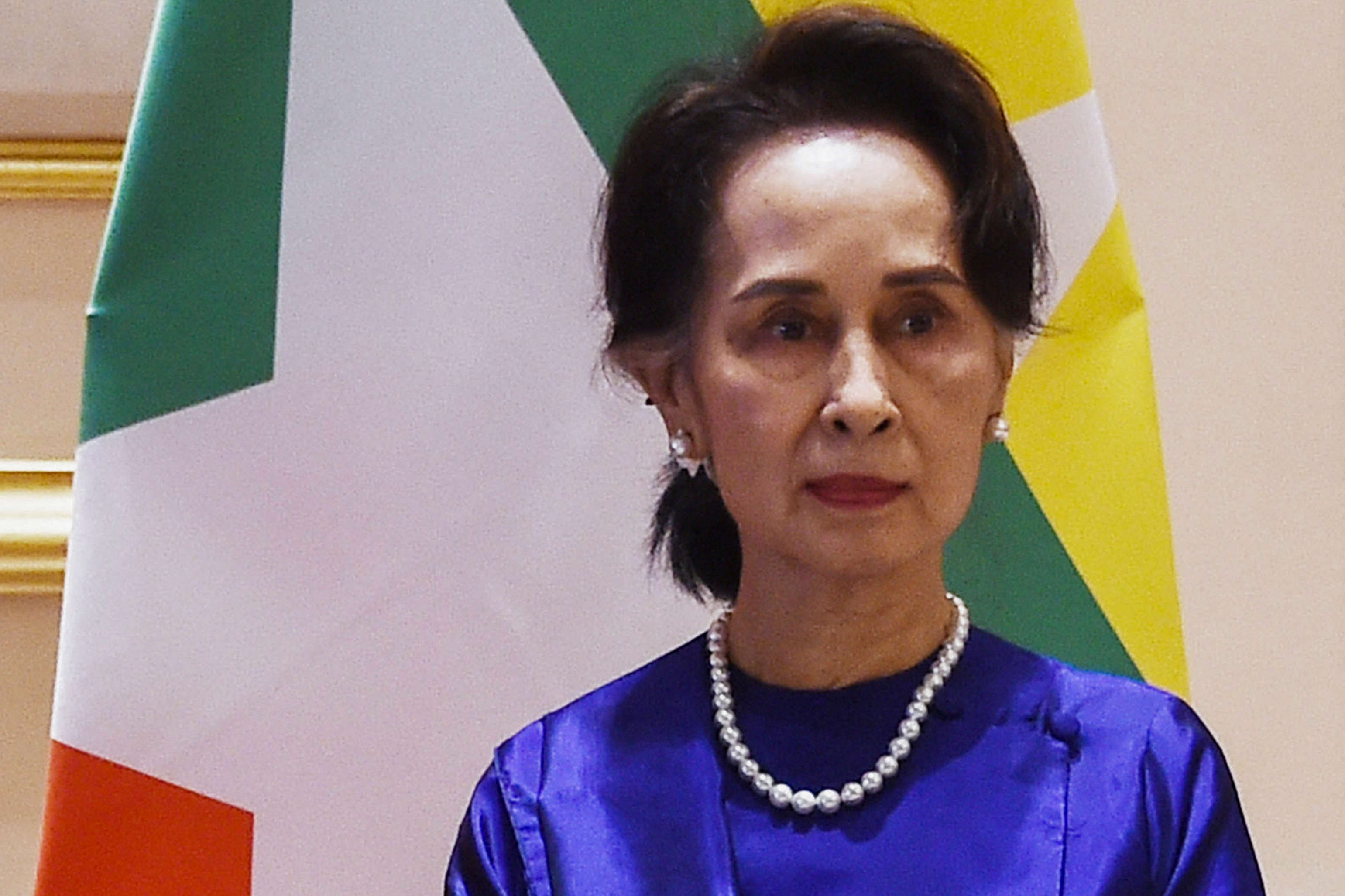 Portrait of Aung San Suu Kyi in royal blue top and string of pearls in front of a Myanmar flag