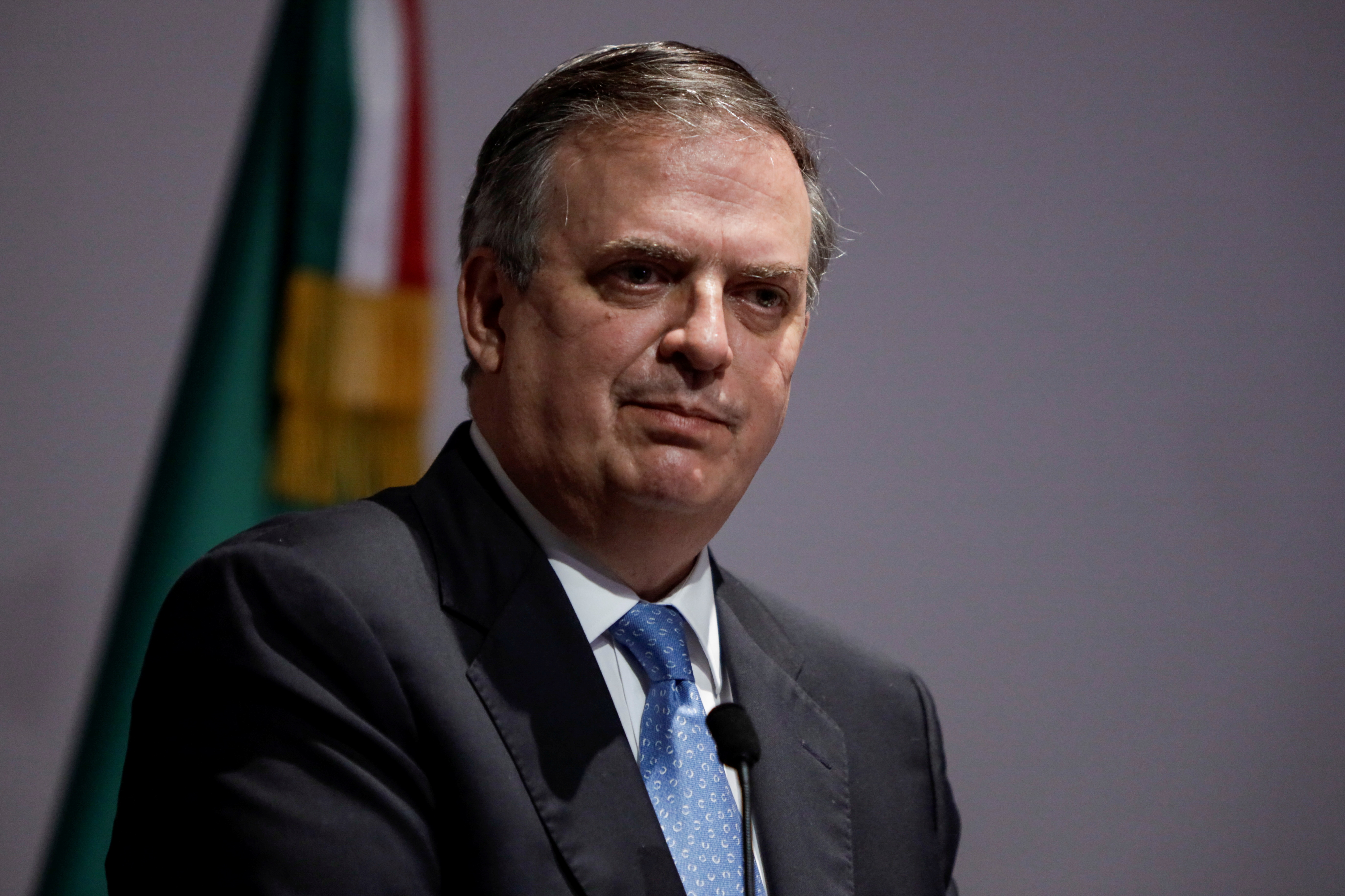 Marcelo Ebrard, in suit, against grey background with some flag showing.