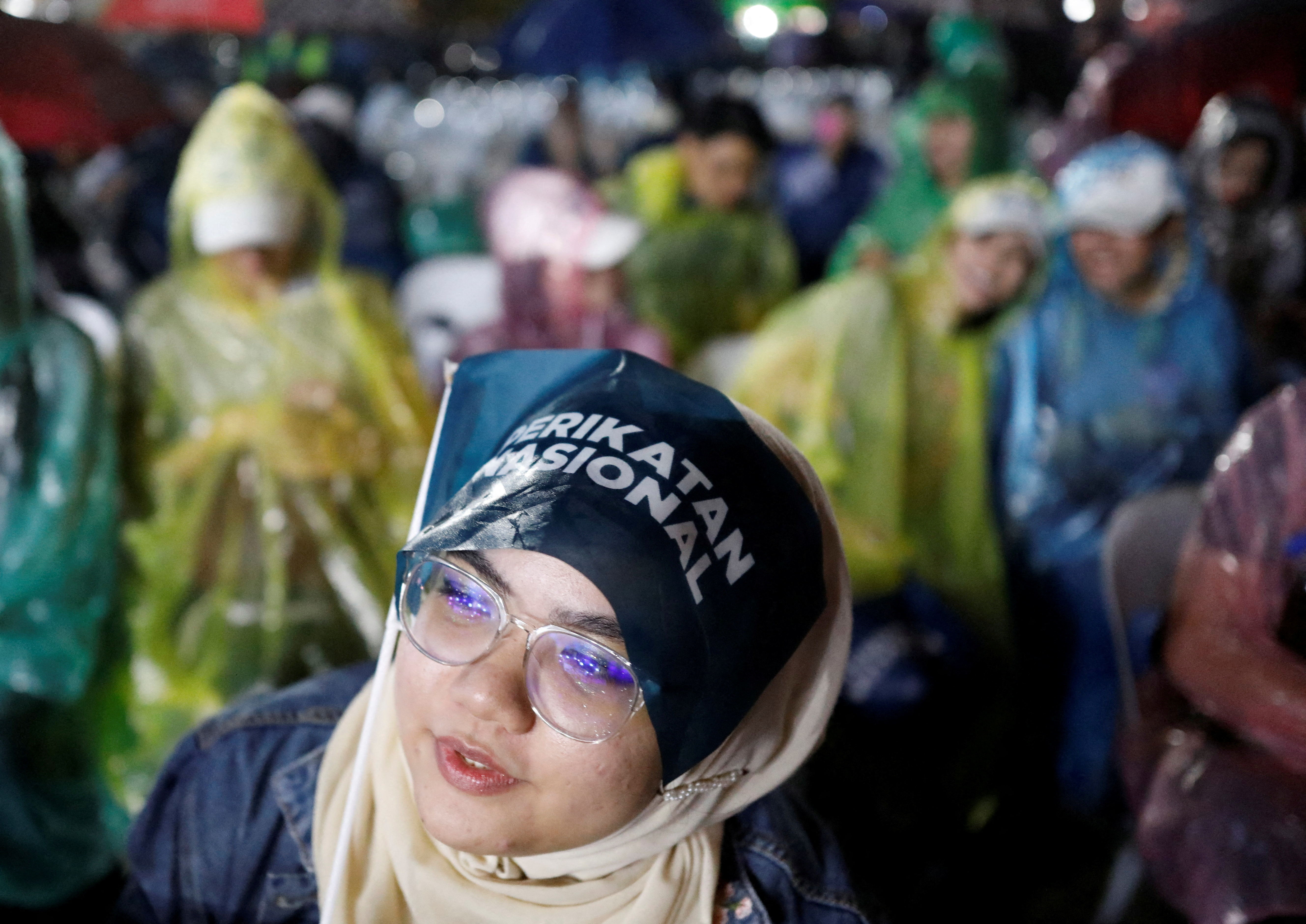 A female supporter of Perikatan Nasional at a rally. She looks to be listening intently. People behind her are wearing raincoats
