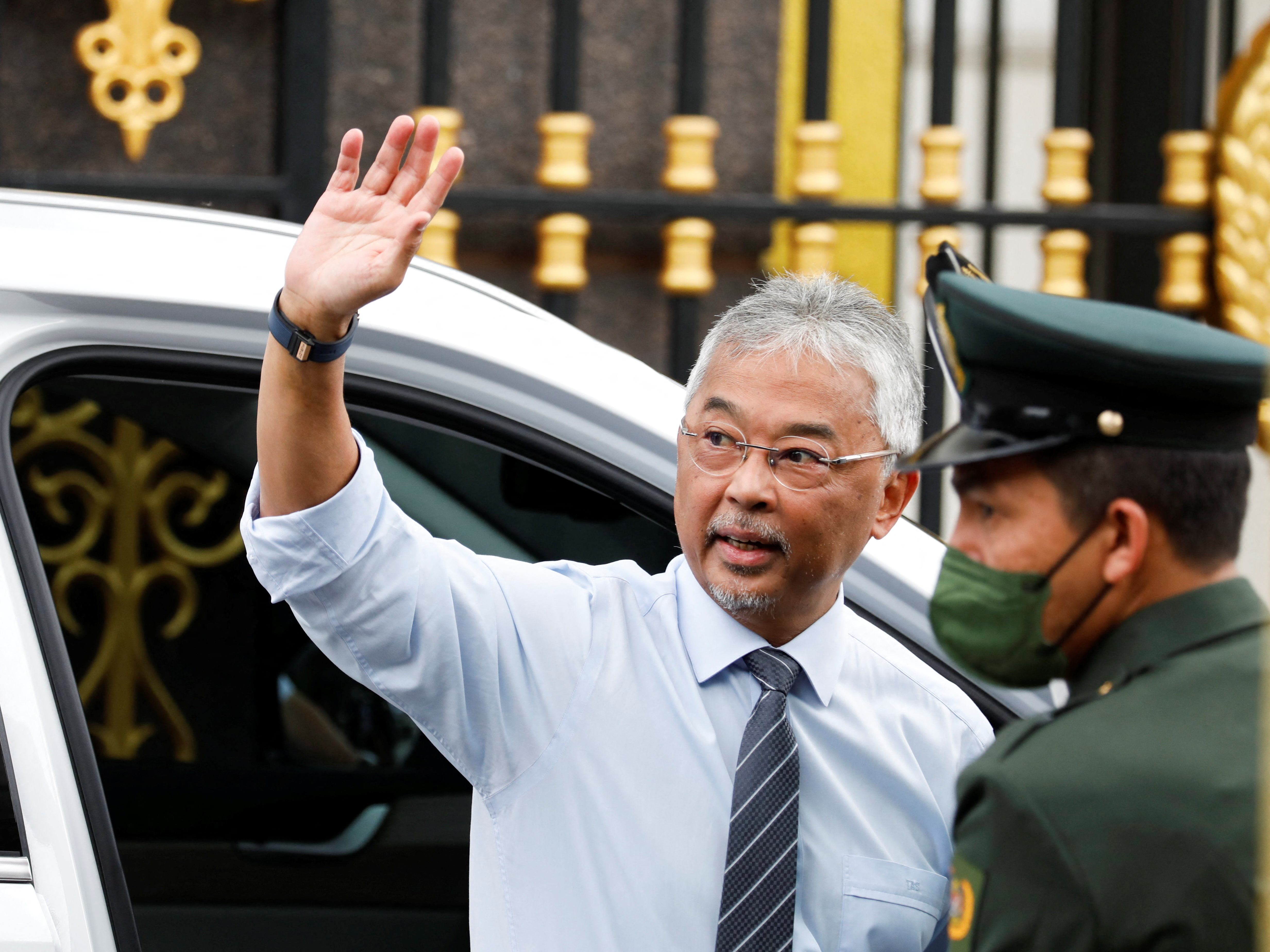 The king in a white shirt and striped tie waves to reporters gathered outside the palace.