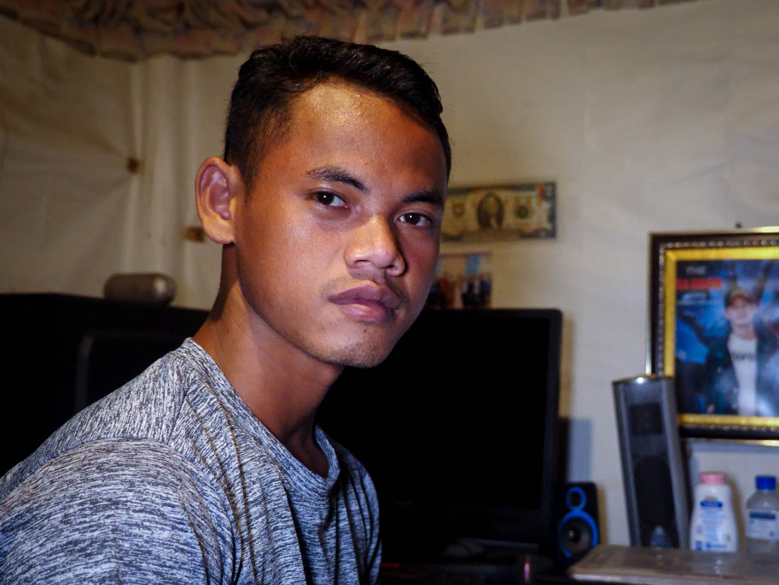 Kea Sokun at his home after he was released from prison. He is wearing a grey t-shirt and looks pensive. There are family photos behind him.