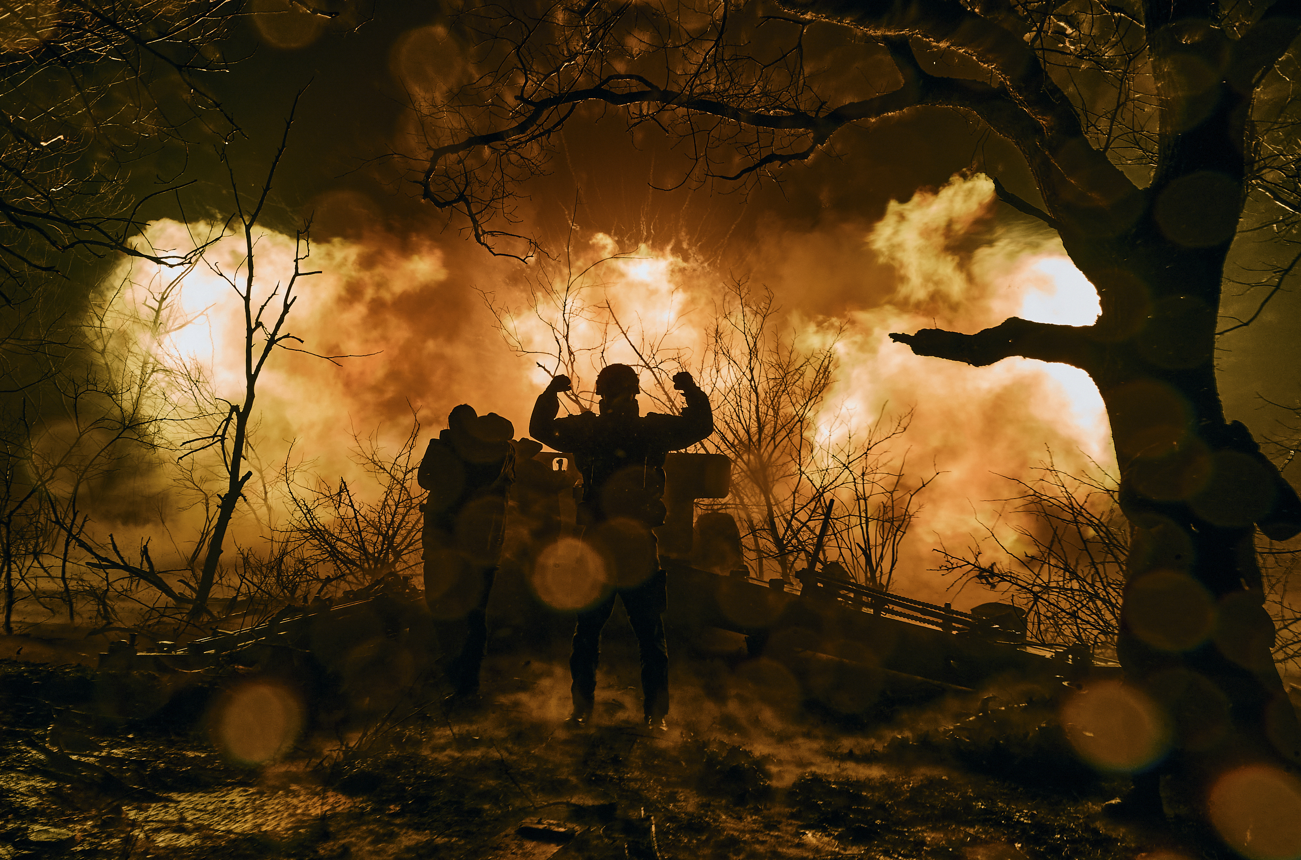 A Ukrainian soldier silhouetted against orange flames after firing artillery at Russian positions near Bakhmut, Ukraine. He has his arms in the air. There are trees at the sides in the foreground and debris behind.