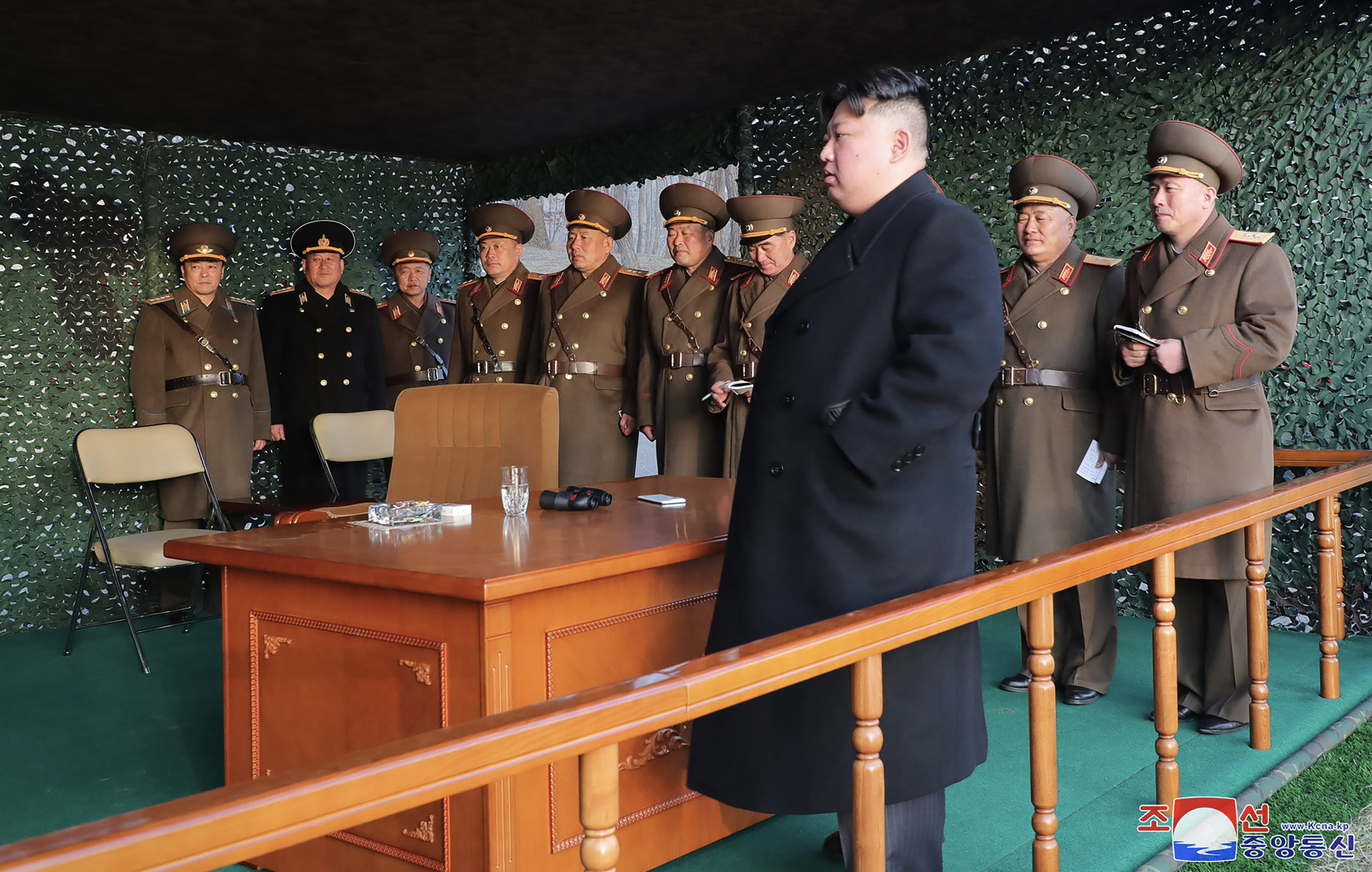 Kim Jong Un in a black coat standing next to a wooden desk in a shelter. Behind him are a row of military officers in uniform.
