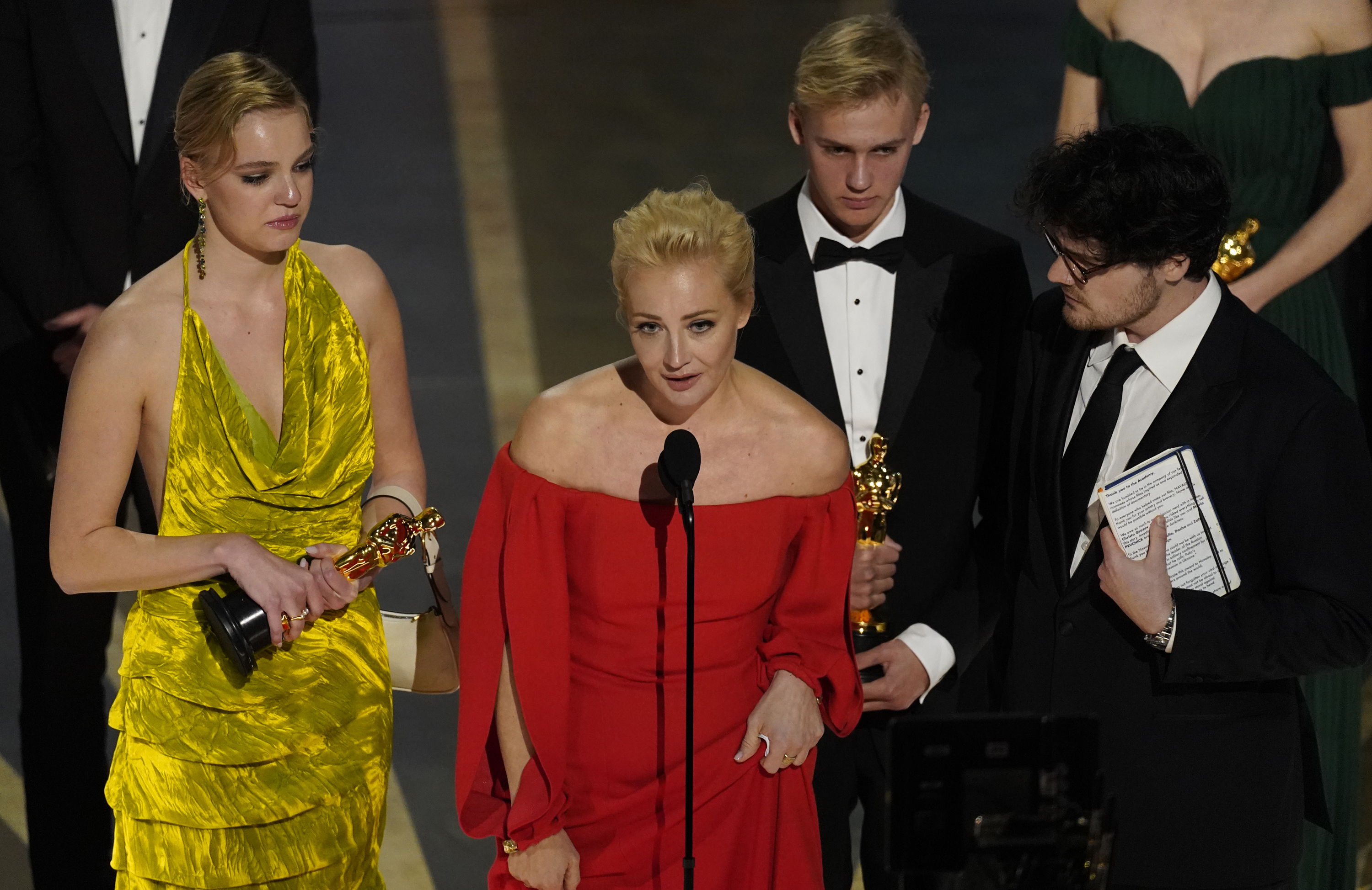 A woman in a red dress speaks into a microphone, surrounded by filmmakers