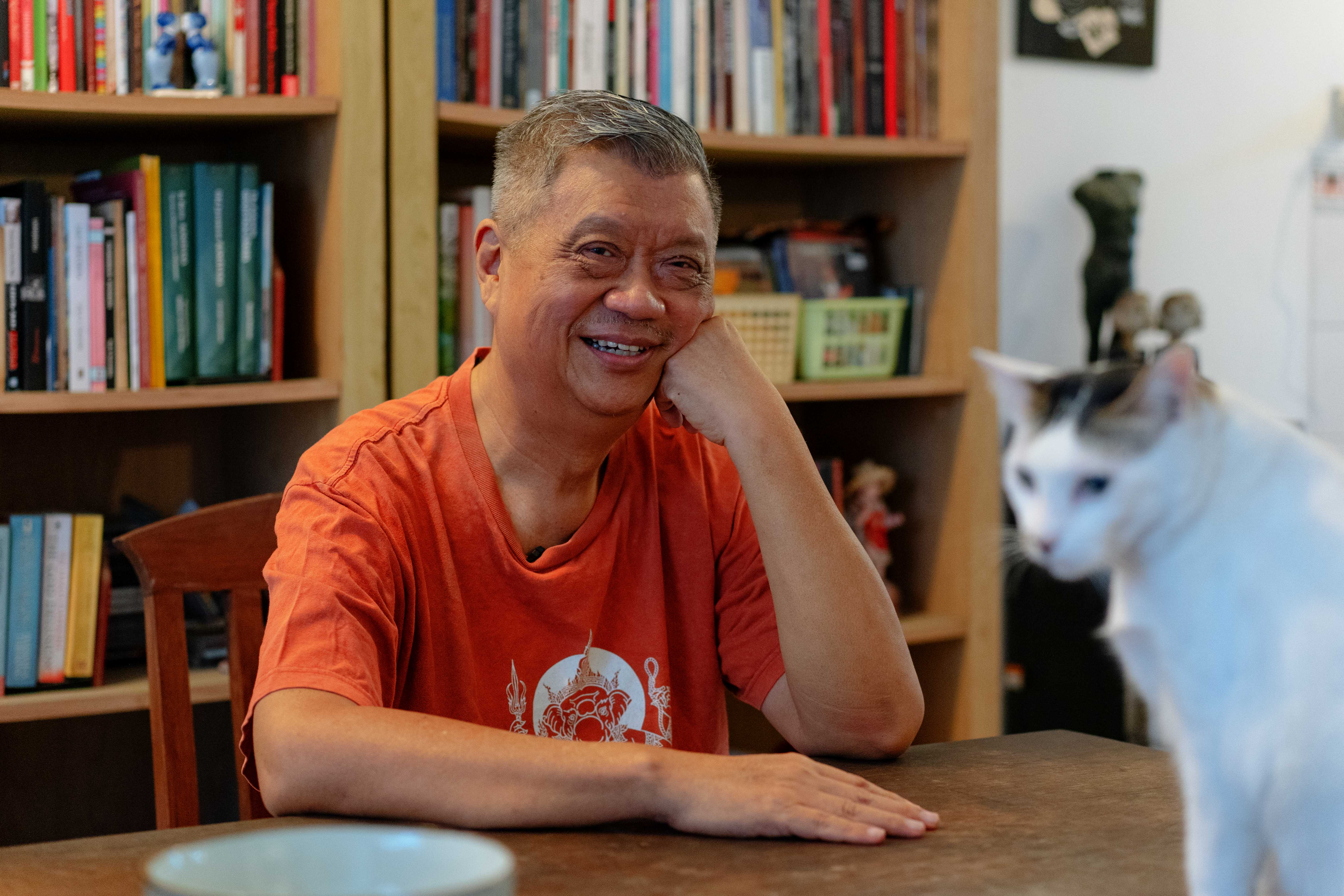 Dédé Oetomo sitting at a desk with a book case behind him. He is smiling and propping his head up on one hand. There's a calico cat walking on the desk in front.