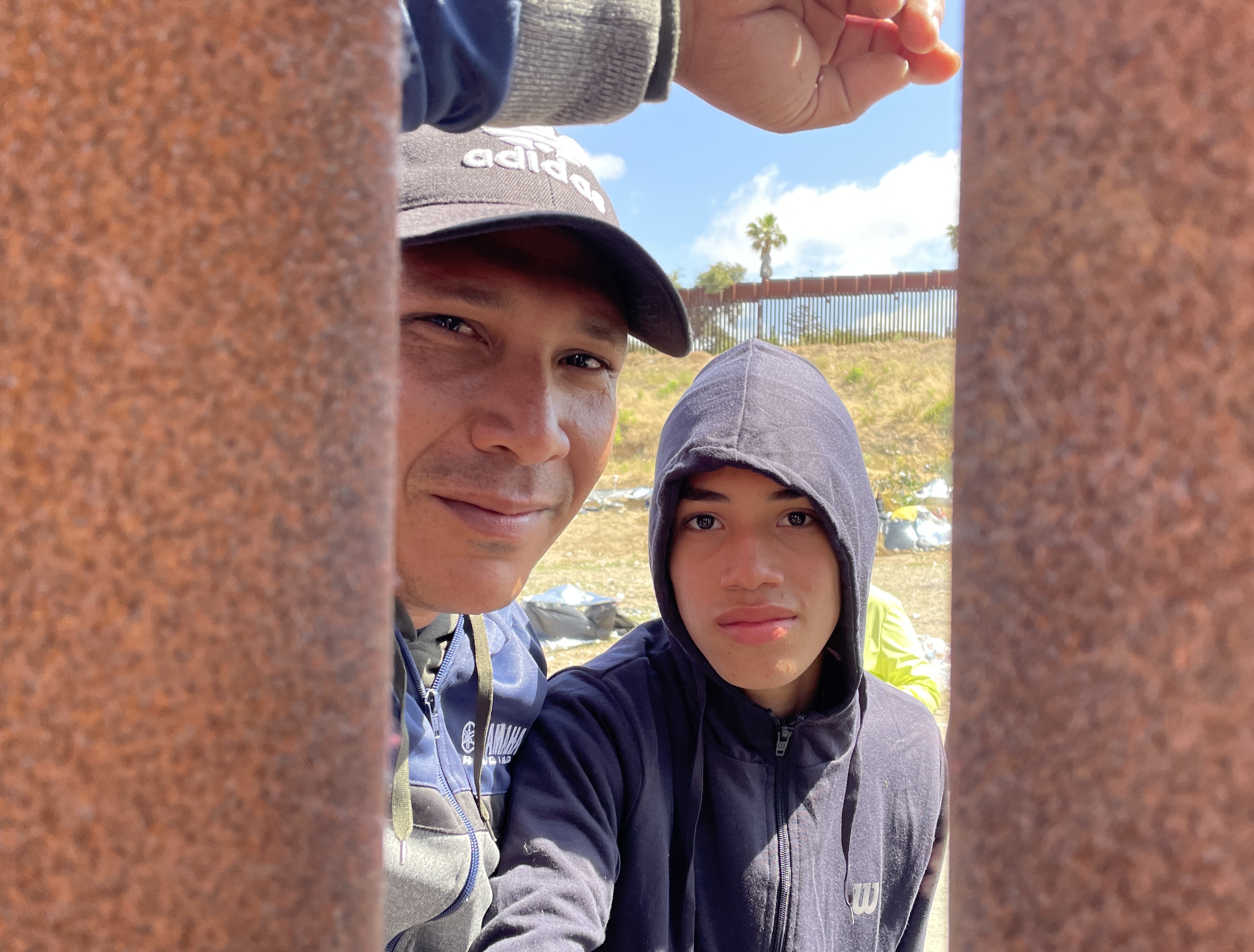A man leans against a metal wall and peers through the gaps between its slats with his son, who wears a hoodie