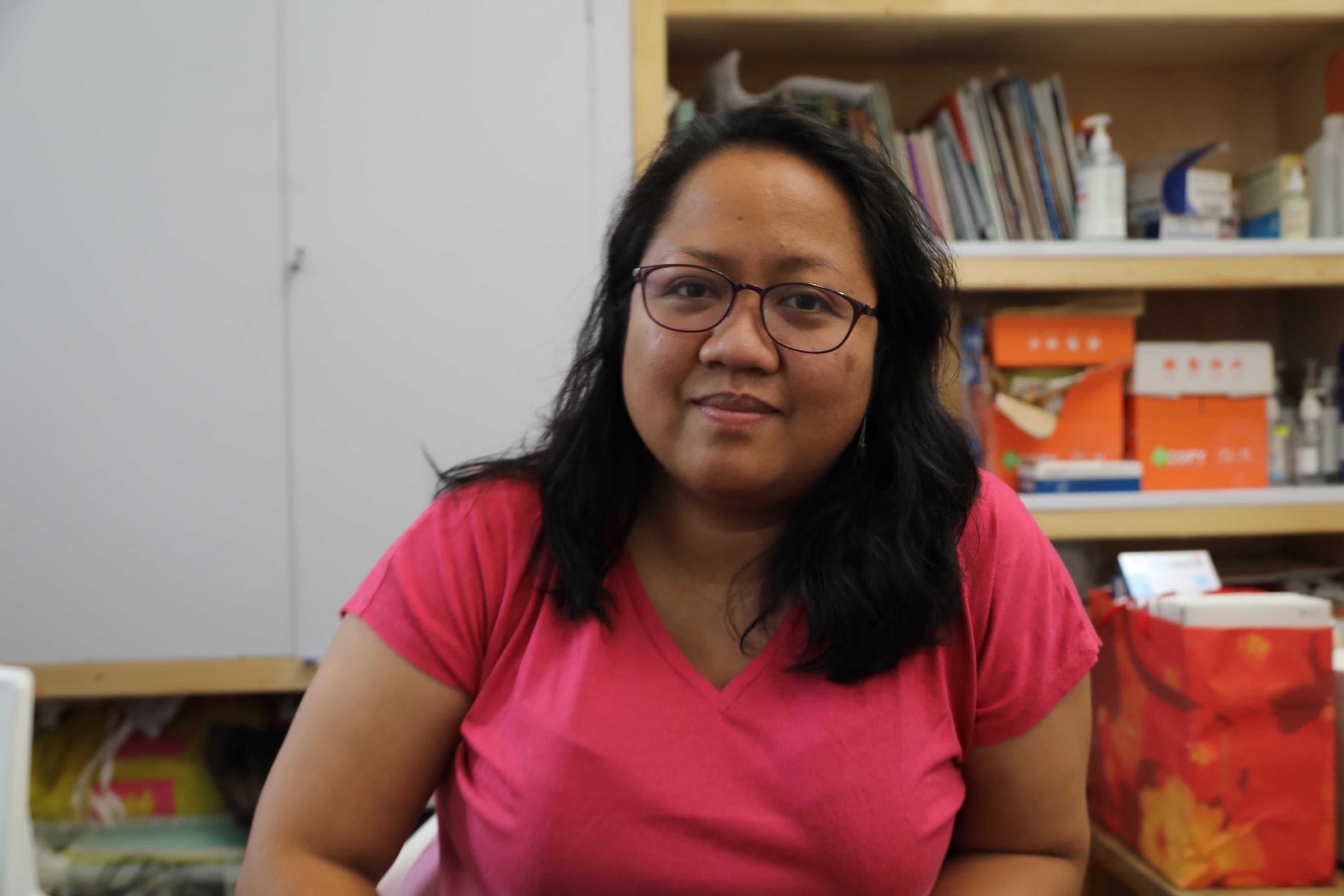 A portrait of Shiela Tebia-Bonifacio. She is sitting  at a desk with a bookcase behind her.