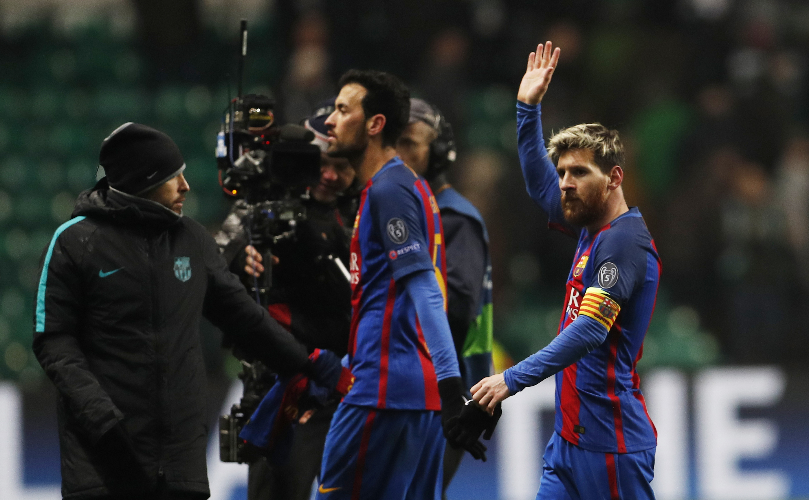 Two men walk across a field as cameras follow them, both dressed in the Barcelona soccer jersey: blue and red stripes. One raises his hand to wave.
