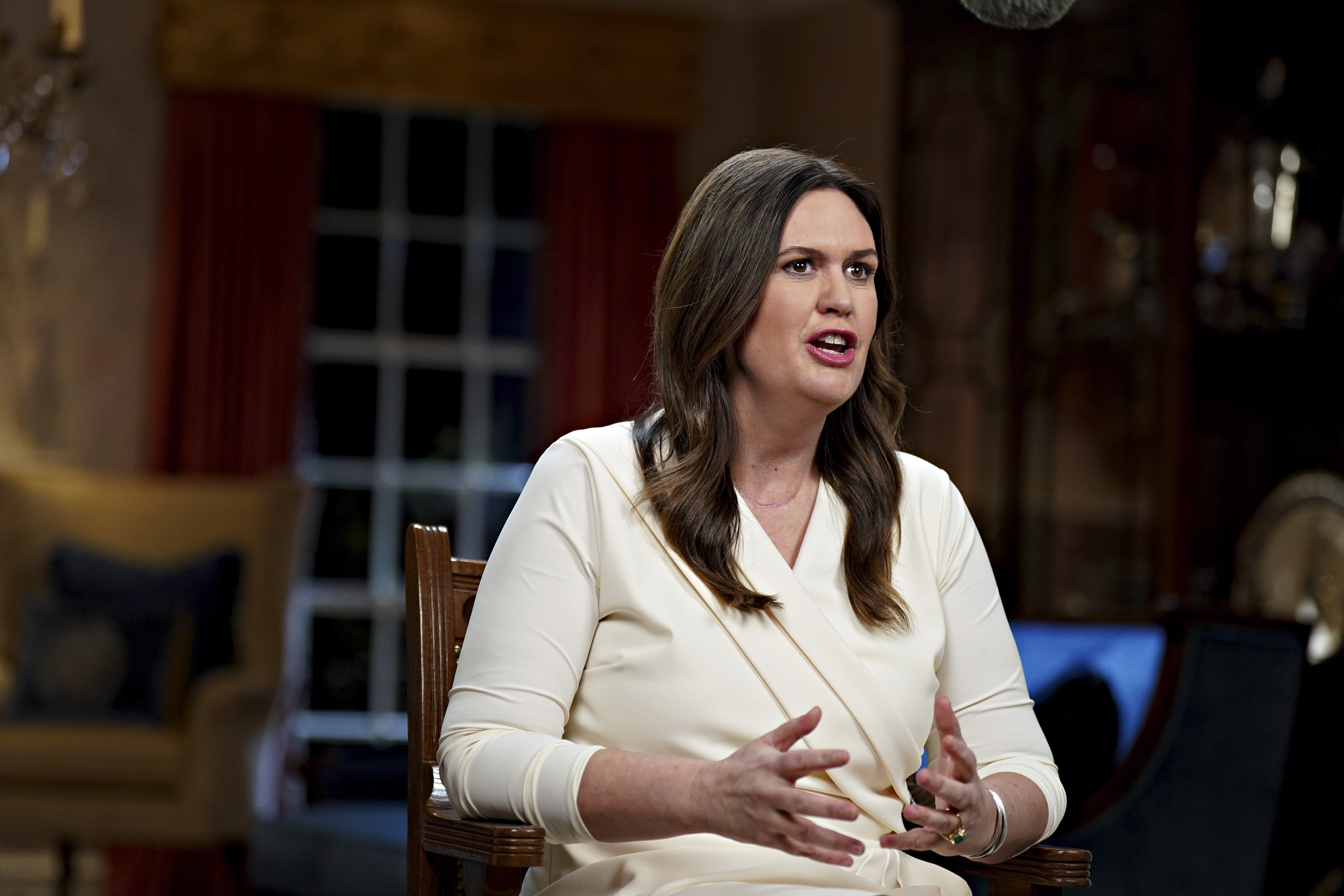 A woman in a white suit speaks while seated in a wooden chair.