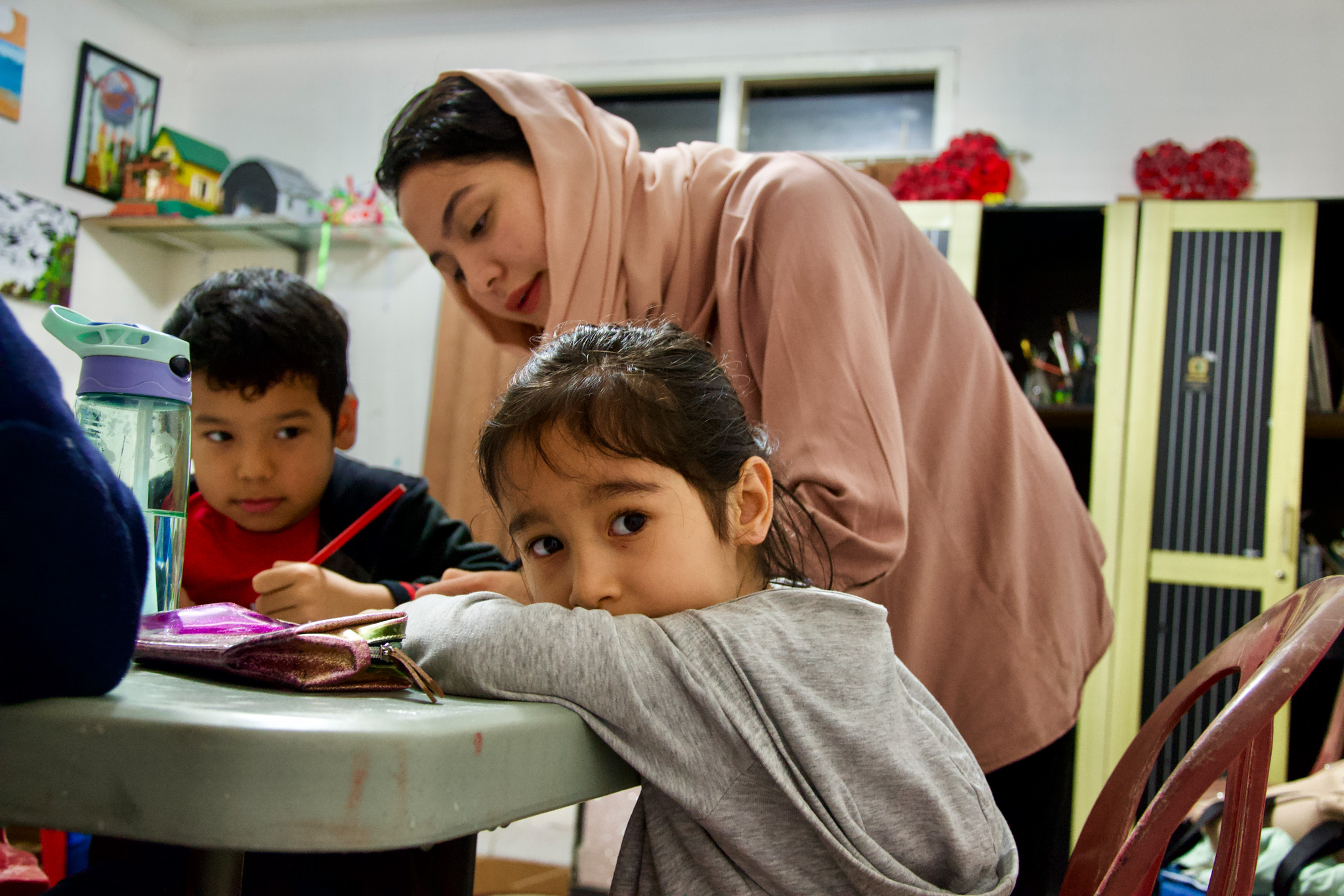 An Afghan women teaching refugee children in Indonesia. One child has turned to look at the camera with their elbows over their books. The teacher is wearing a pale pink outfit with a headscarf tied loosely around her dark hair.