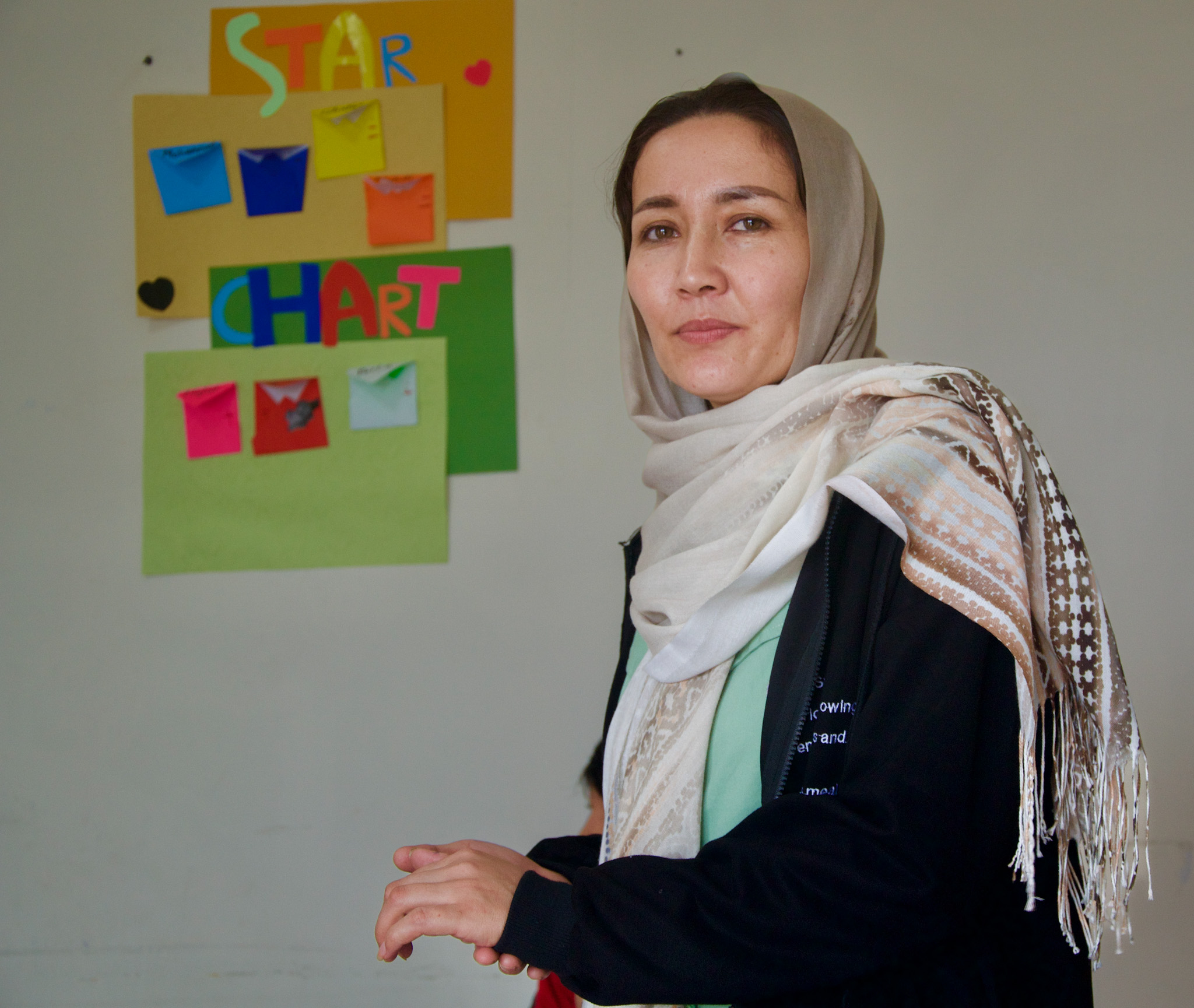 Sofie in a classroom. She is wearing a black top and has draped a pale coloured shawl around her head and shoulders. There is a colourful 'Star Chart ' on the wall behind her