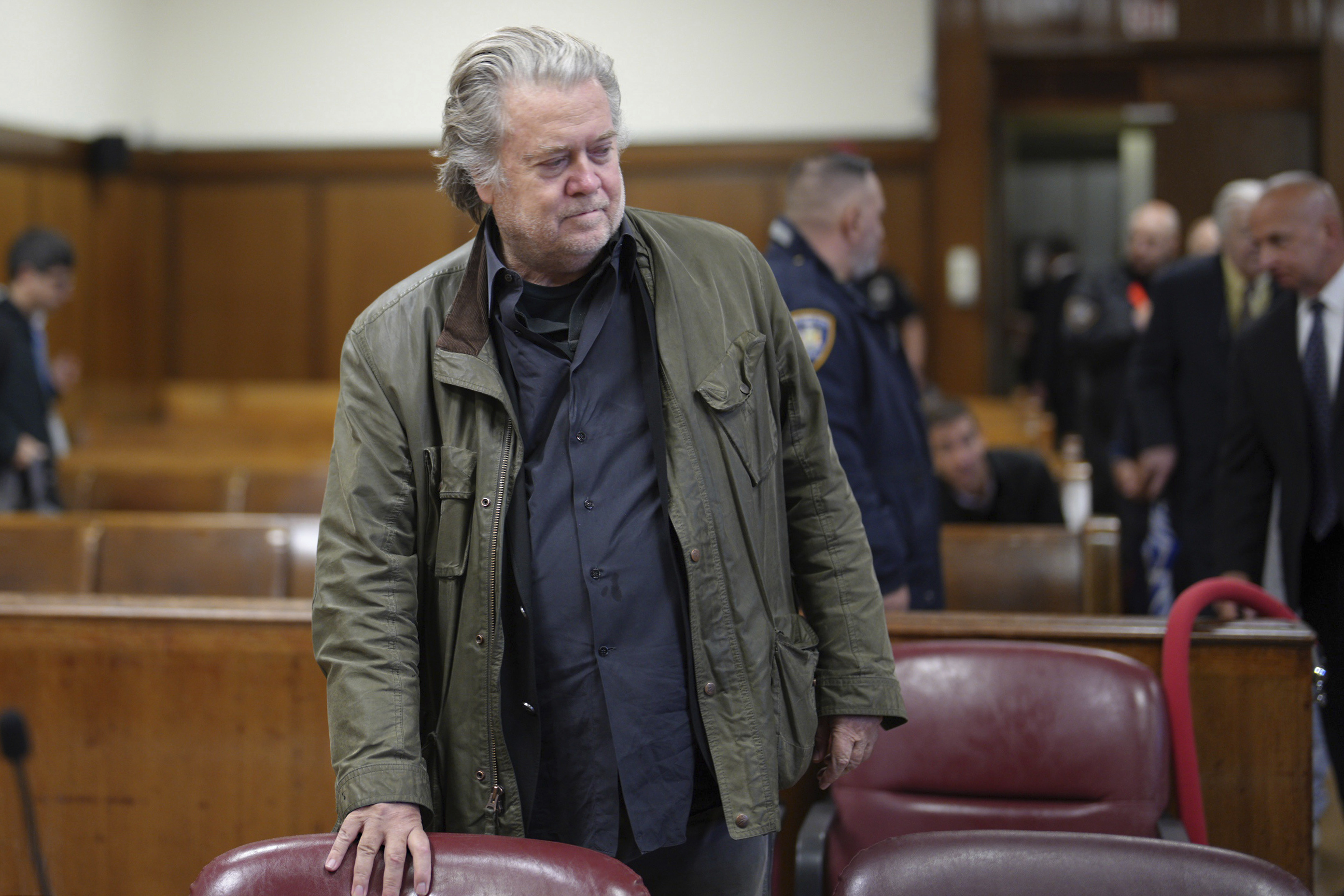 A man in a blue collared shirt and an olive green jacket stands in a courtroom, one hand on a seat.