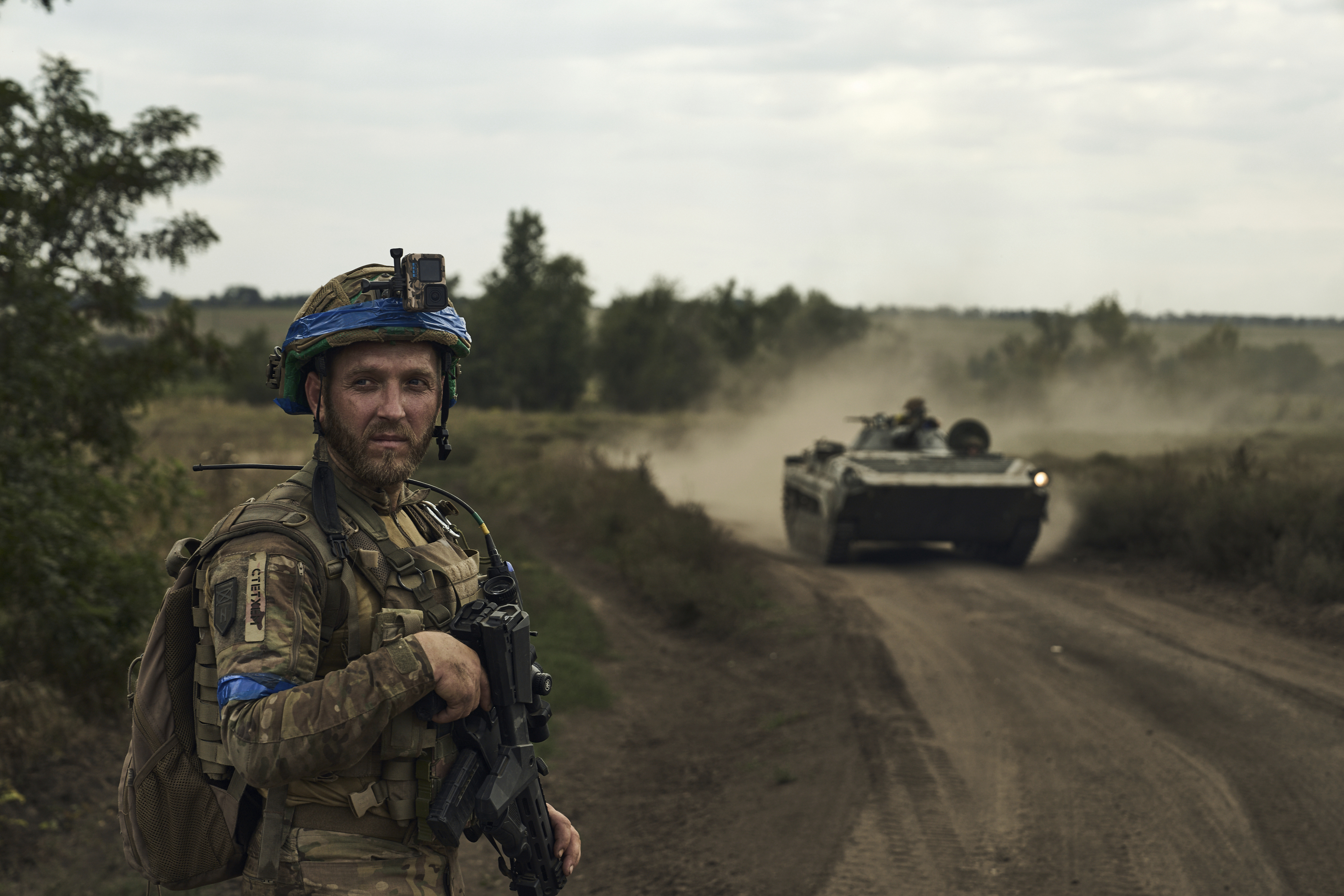 A soldier stands by the side of a dirt track. A tank is advancing along the road in a cloud of dust