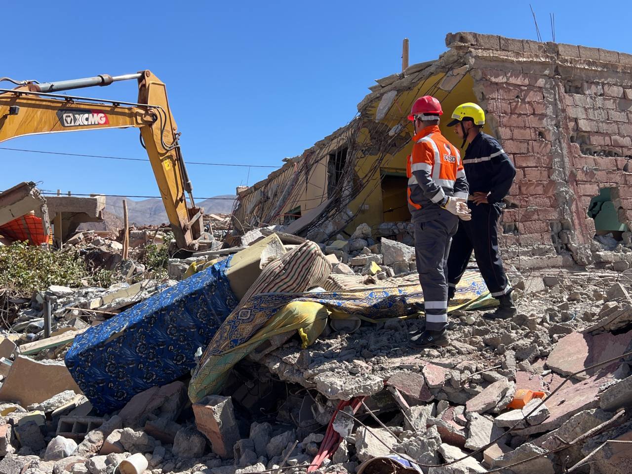 Rescuers stand on rubble watching a backhoe