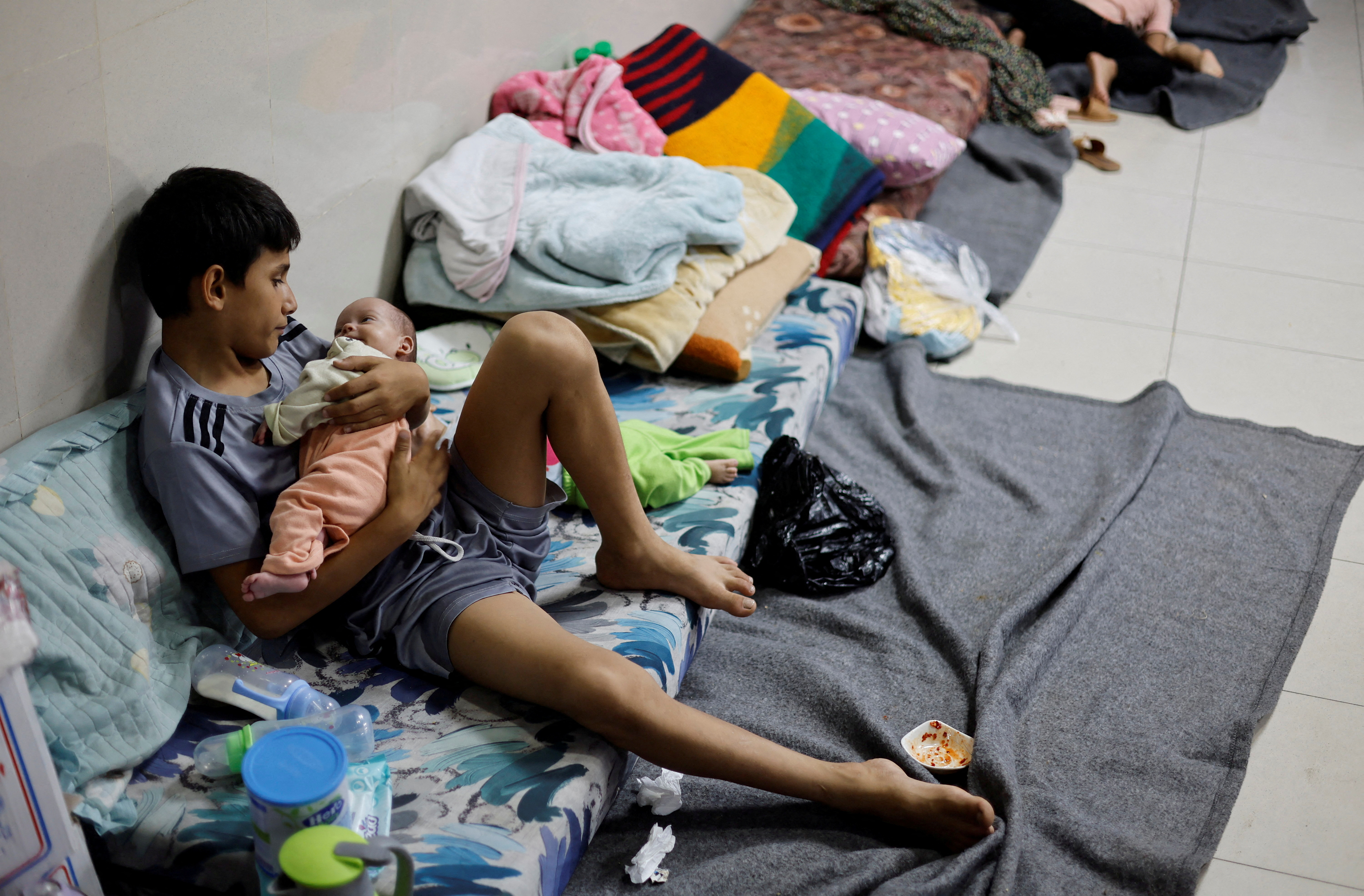 A boy cradles a little baby in his arms as he lies on blankets on the floors of a hospital.