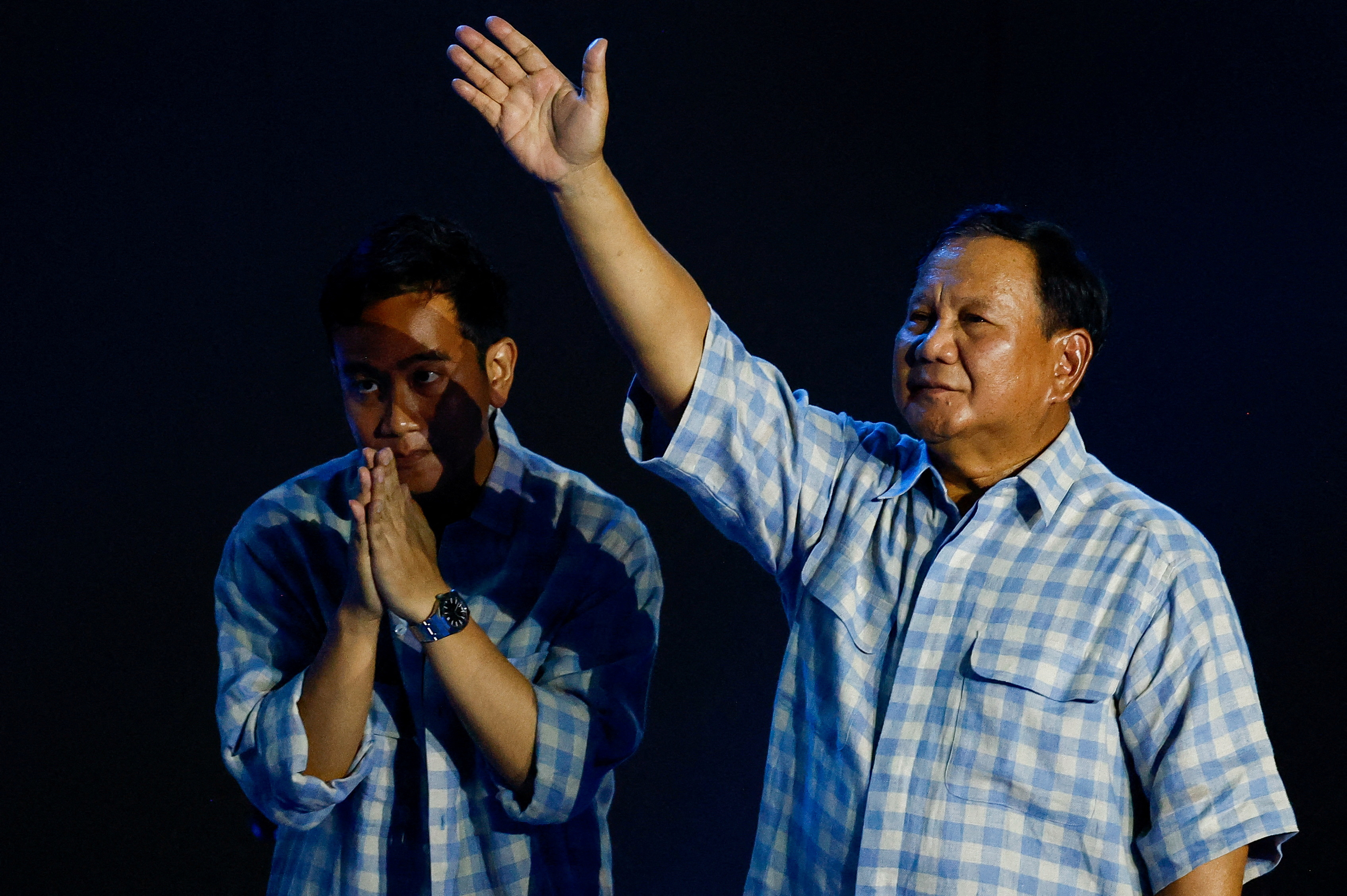 Prabowo Subianto acknowledging his supporters. He has his right arm raised. Running mate Gibran Rakabuming Raka is putting his hands together in gratitude and bowing his head