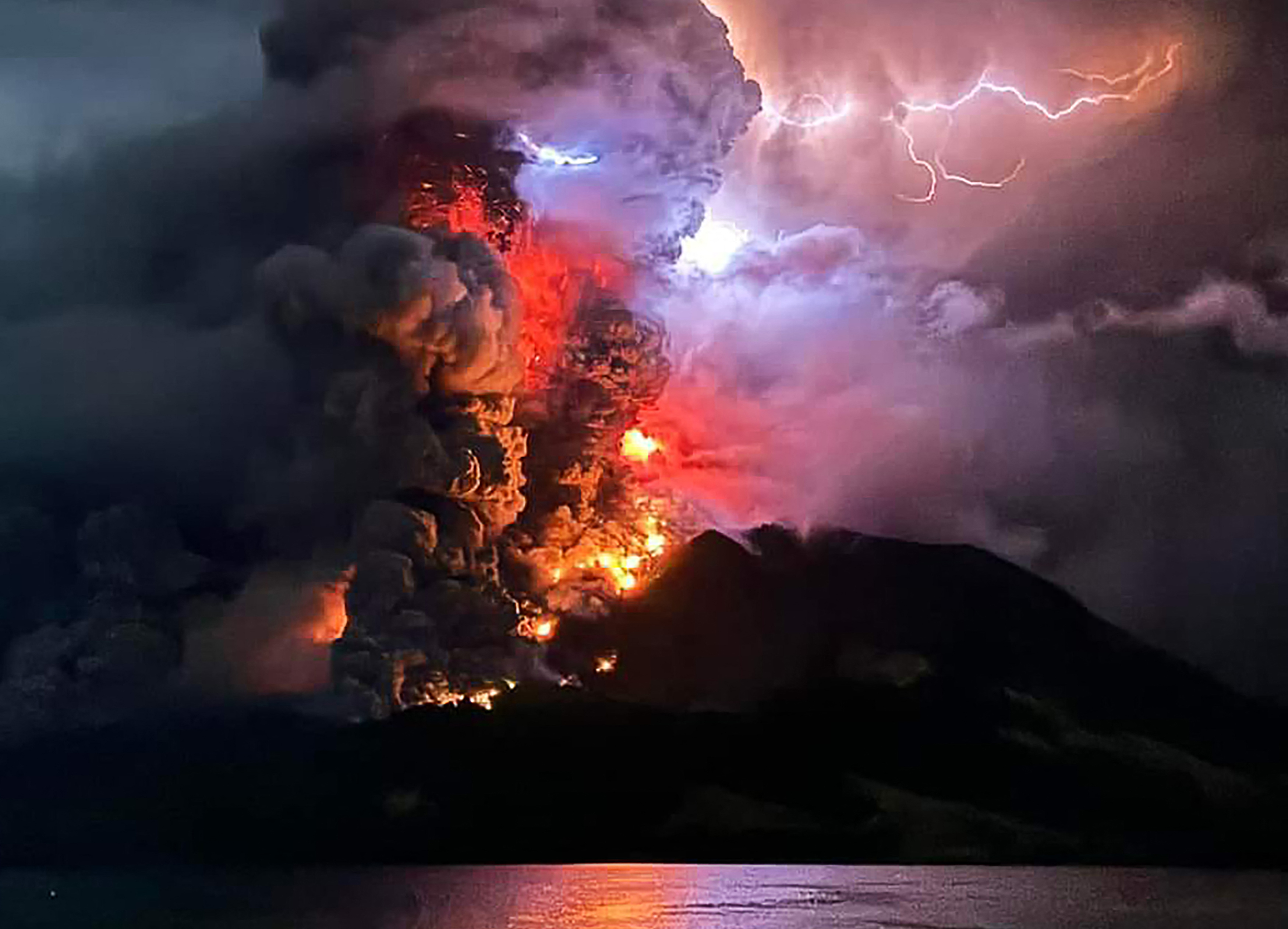 The Ruang volcano erupting. There are billowing clouds of smoke and ash obscuring the sun. Orange flames can also be seen.