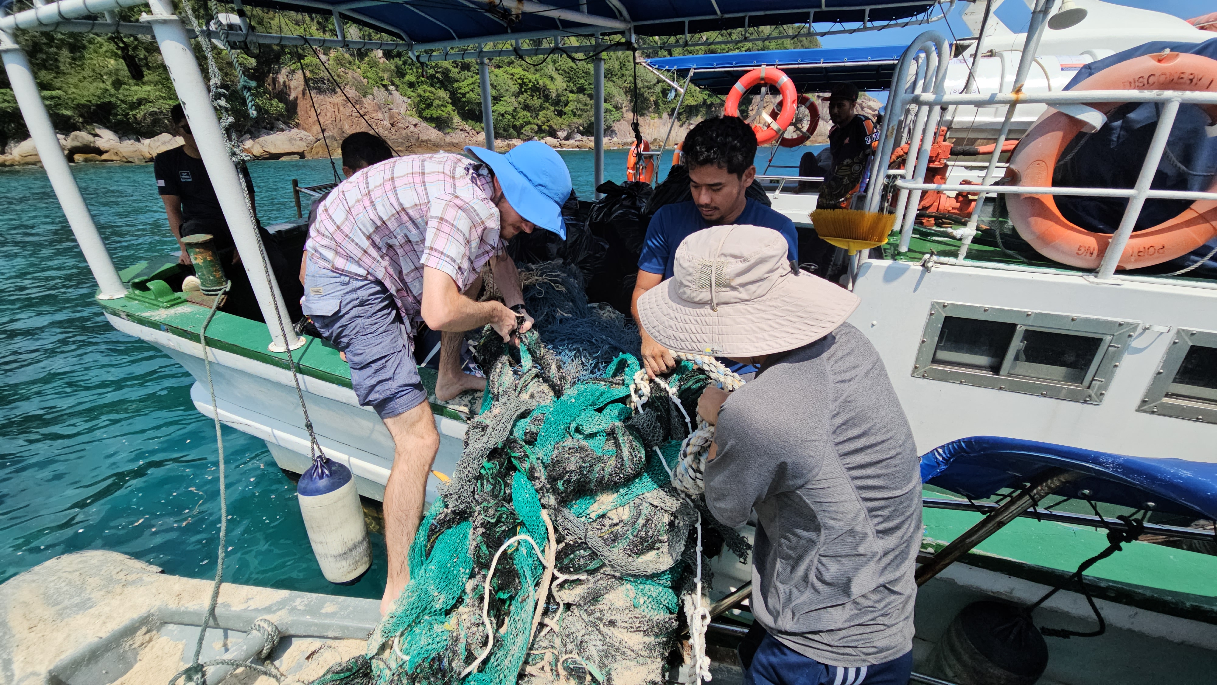 A group of people moving a massive tangle of discarded fishing nets onto a small boat. The nets are green and grey and knotted.