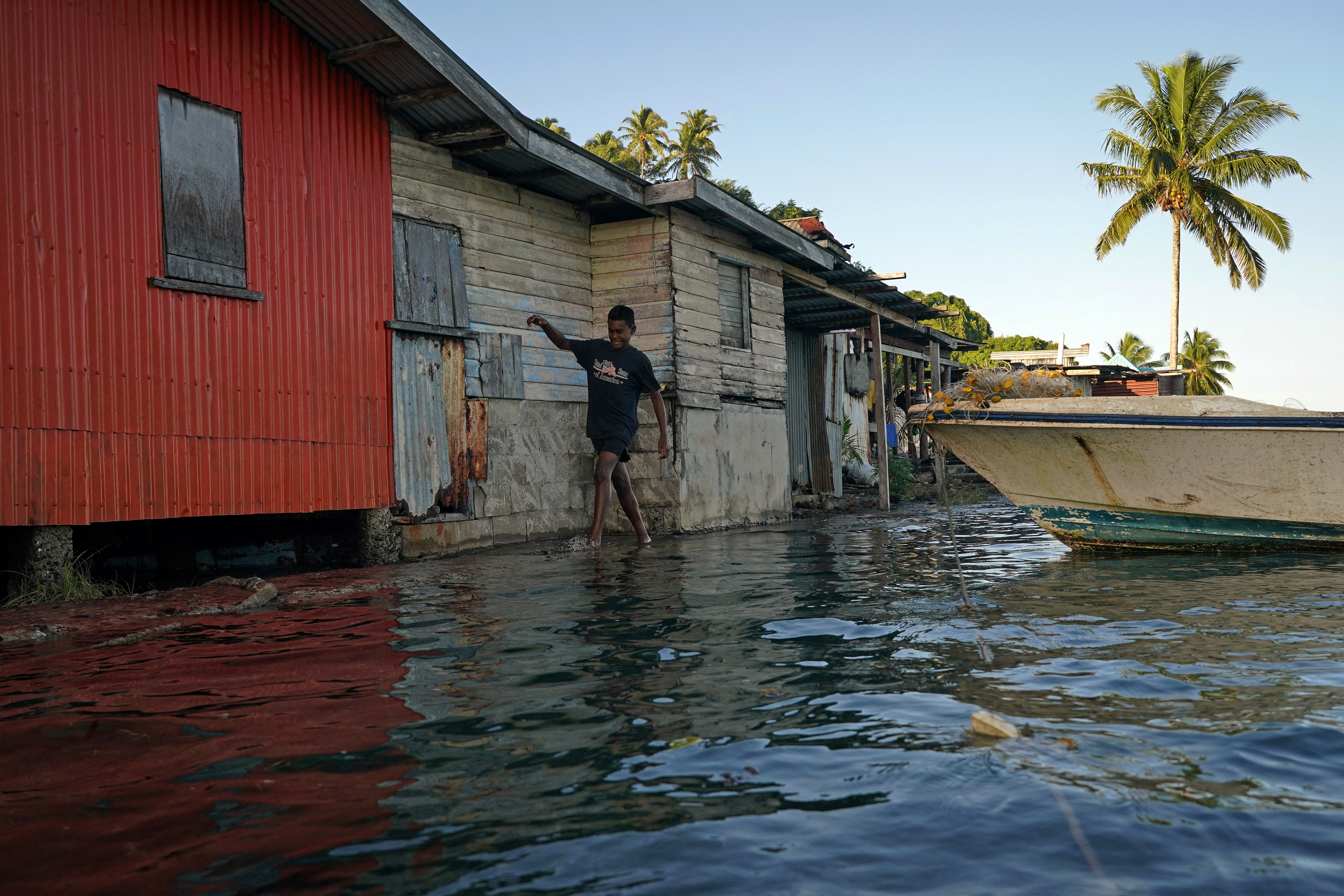 A wood and concrete house in Fiji surrounded by seawater. A boy is wading through the water. A boat is floating nearby. here is a palm tree in the distance