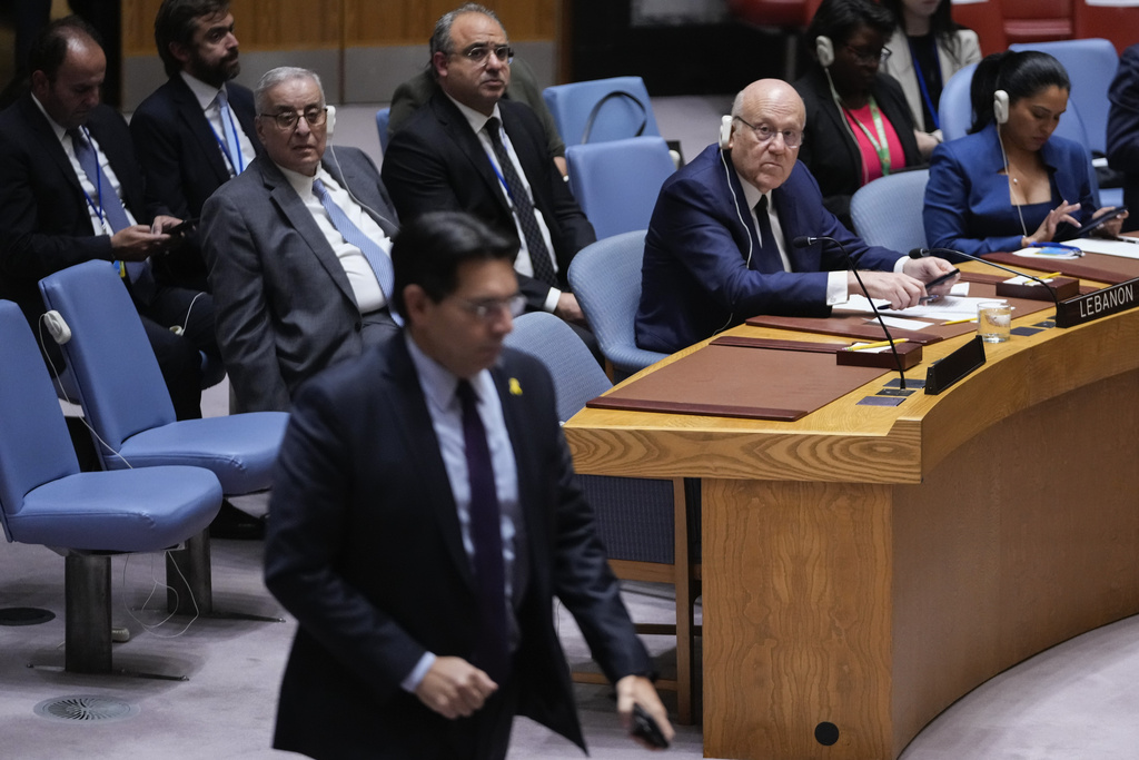 a man sits at a desk that says Lebanon as another man in a suit with a yellow lapel pin walks past him
