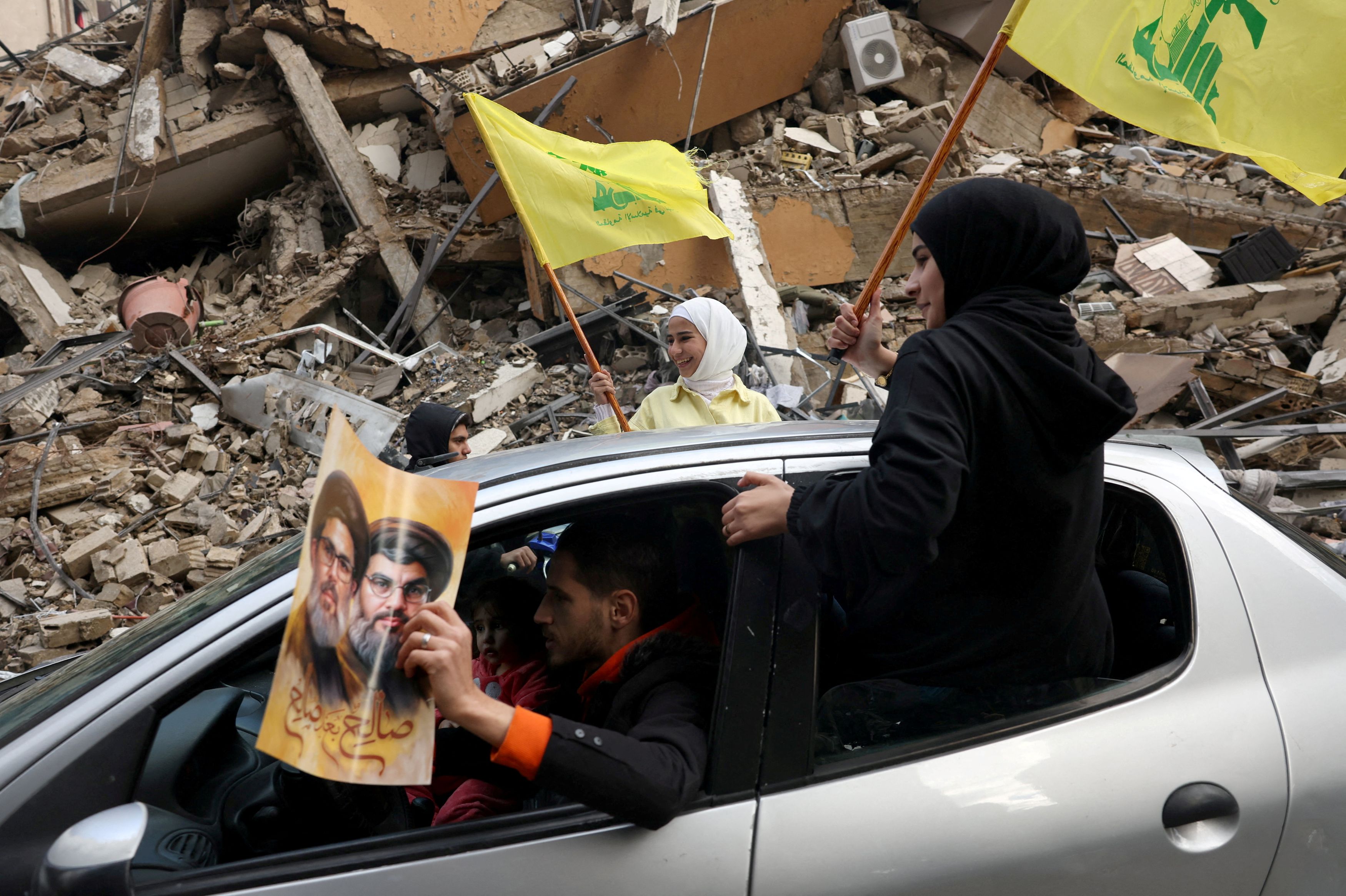 Women wave Hezbollah flags as they drive past a damaged building at the entrance of Dahiyeh