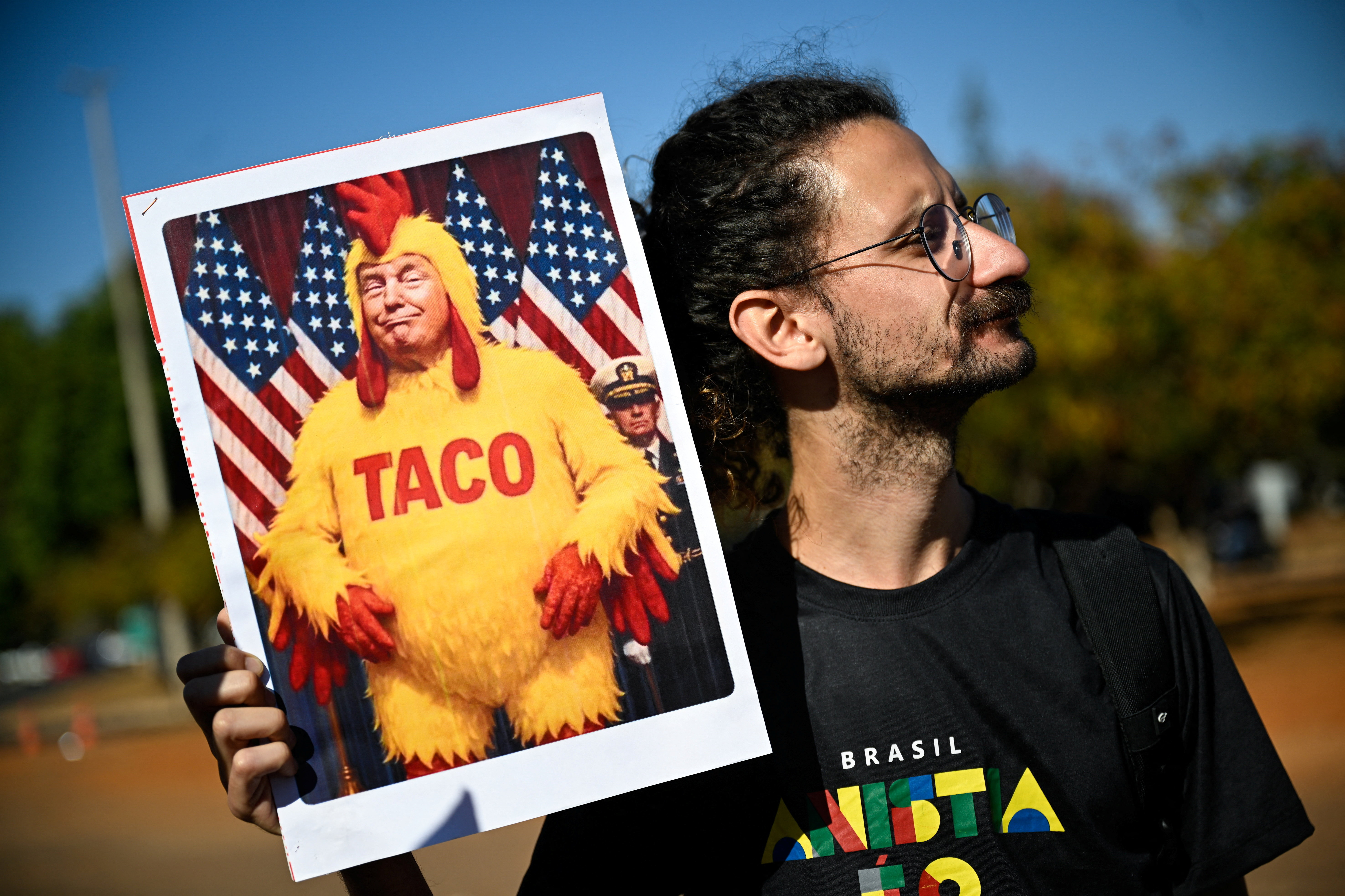 A Brazilian protester holds up a sign with Trump in a chicken suit labelled "TACO"