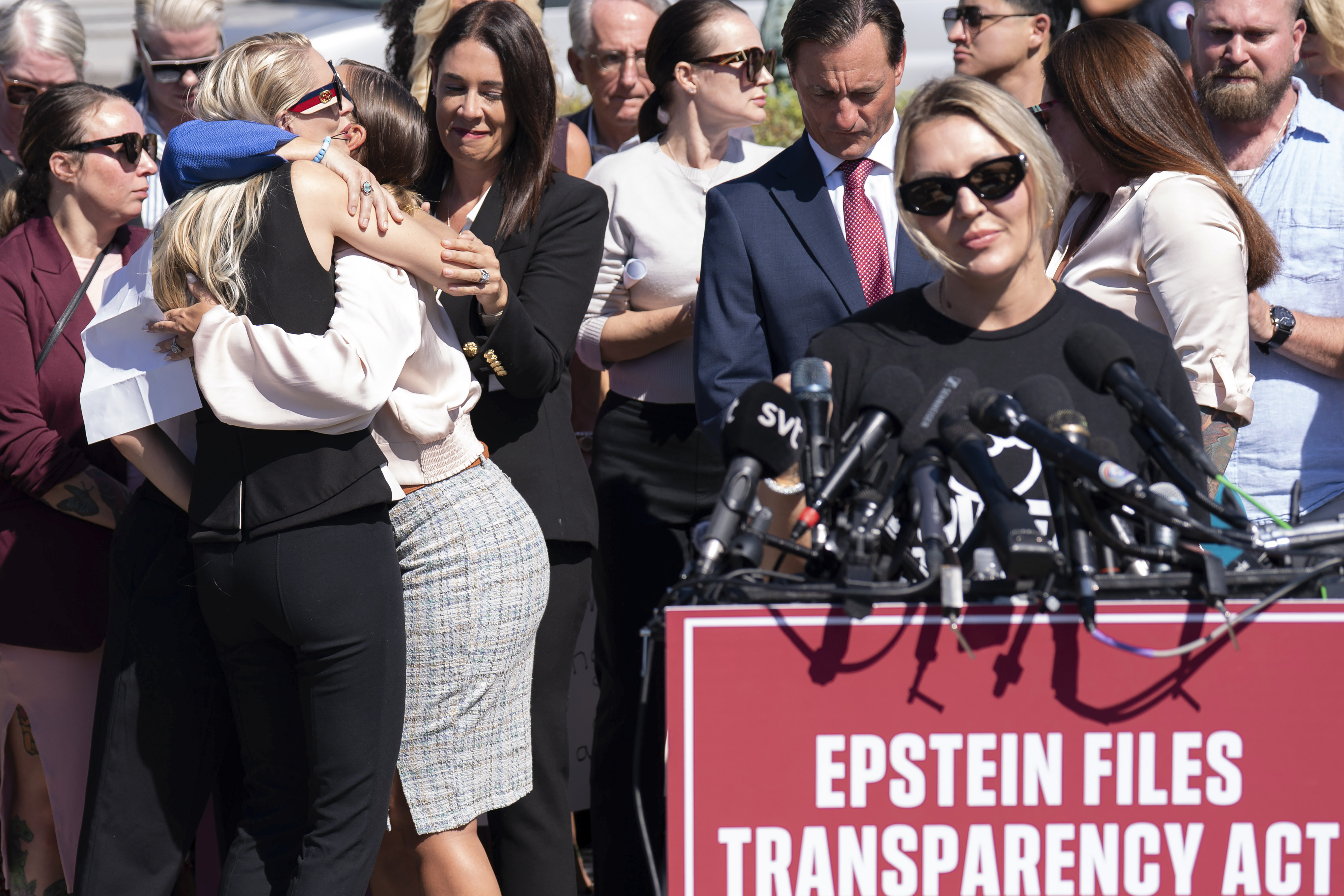 Courtney Wild speaks at a podium while Anouska de Georgiou hugs Marina Lacerda