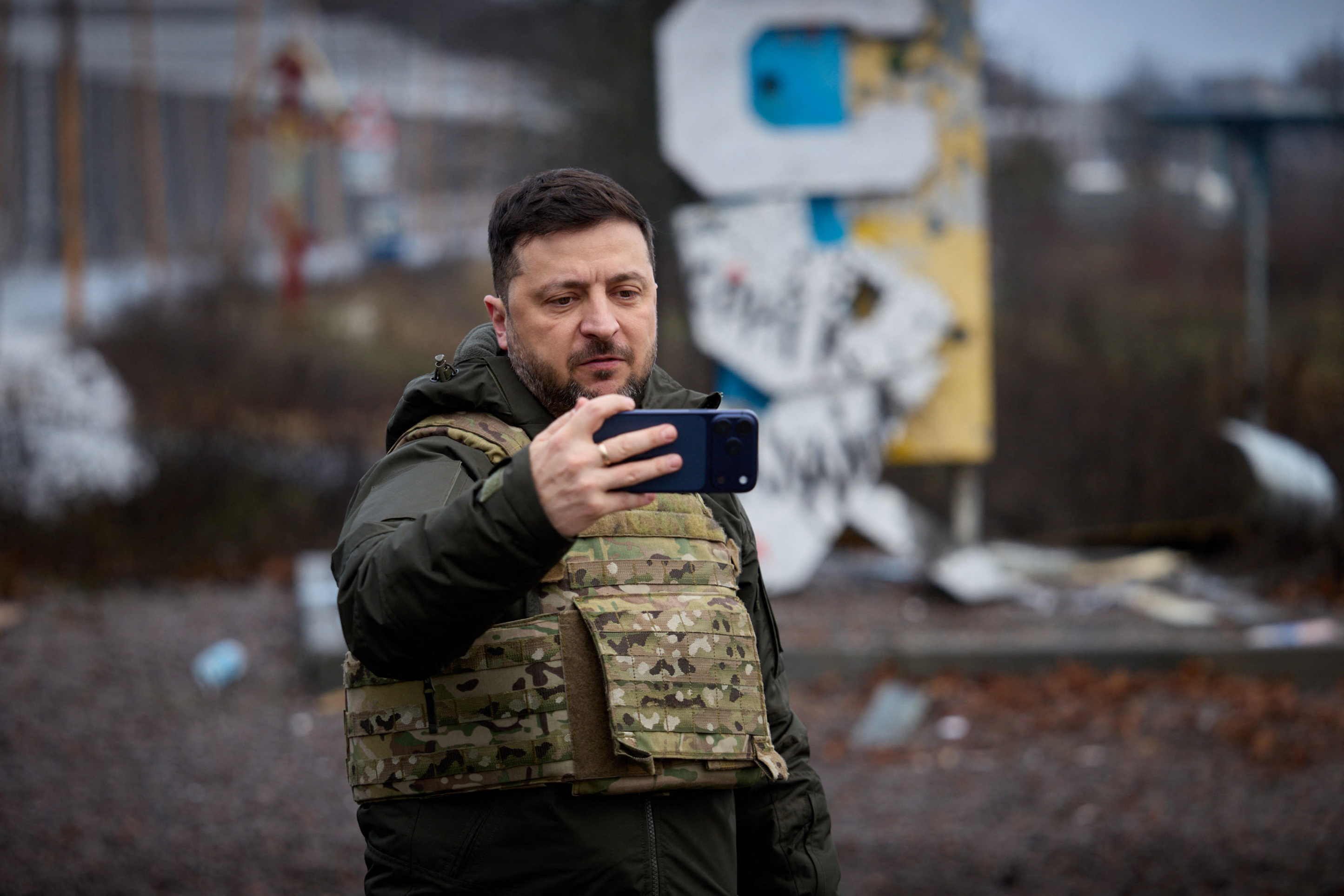 In this handout photograph taken and released by the Ukrainian Presidential Press Service on December 12, 2025, Ukraine's President Volodymyr Zelensky records a video message on his mobile phone in front of the road sign marking the entrance to Kupiansk, Kharkiv region