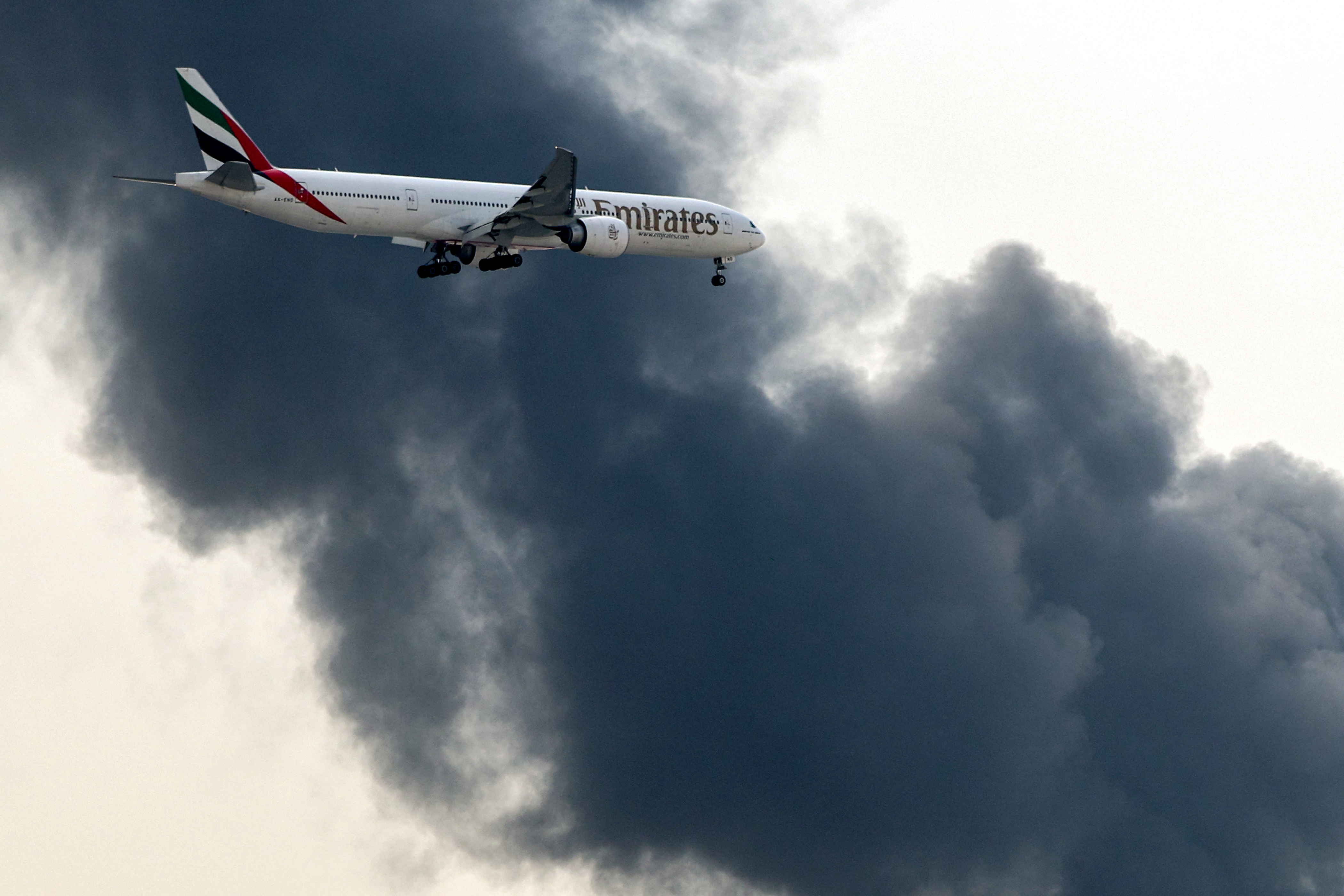 An Emirates Boeing 777 aircraft prepares for landing as a smoke plume rises from an ongoing fire at Dubai International Airport