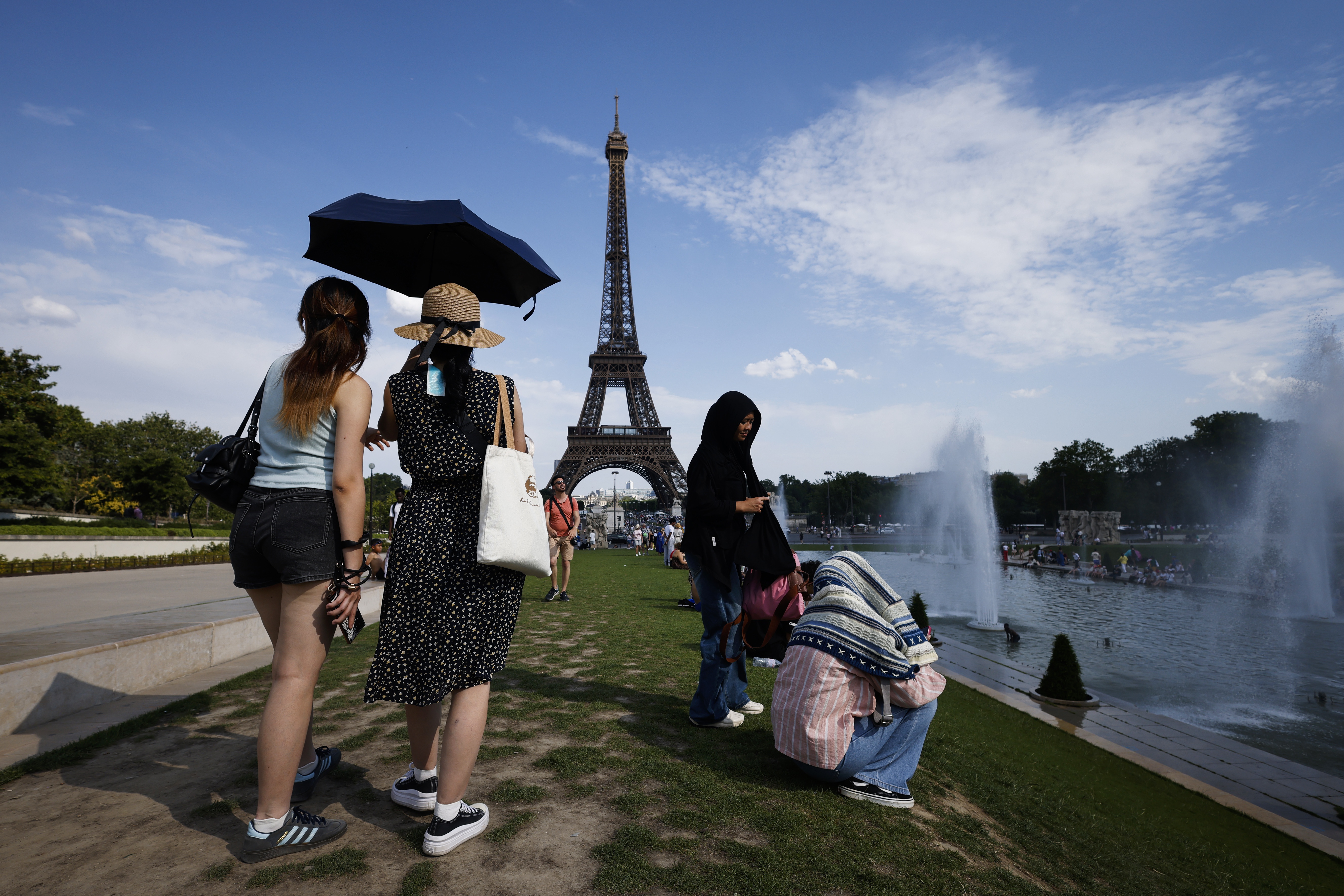 Women shelter from the heat under an umbrella near a fountain, with the Eiffel Tower in the distance.
