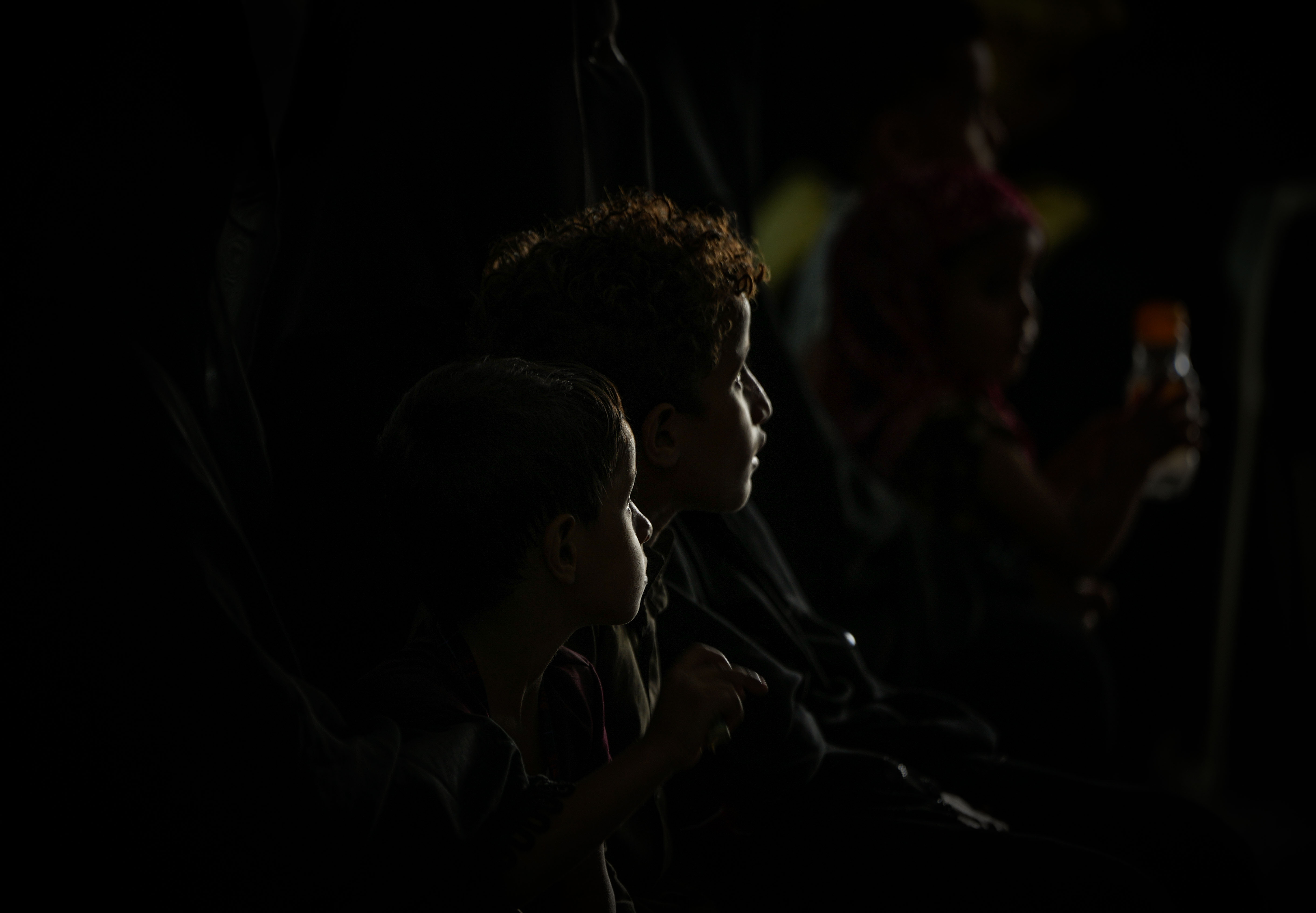 Children sit in darkness, their faces lit by a small screen. During power cuts, some families rely on phones, rechargeable lamps or small solar systems for light at night. Photo: Belal Khaled