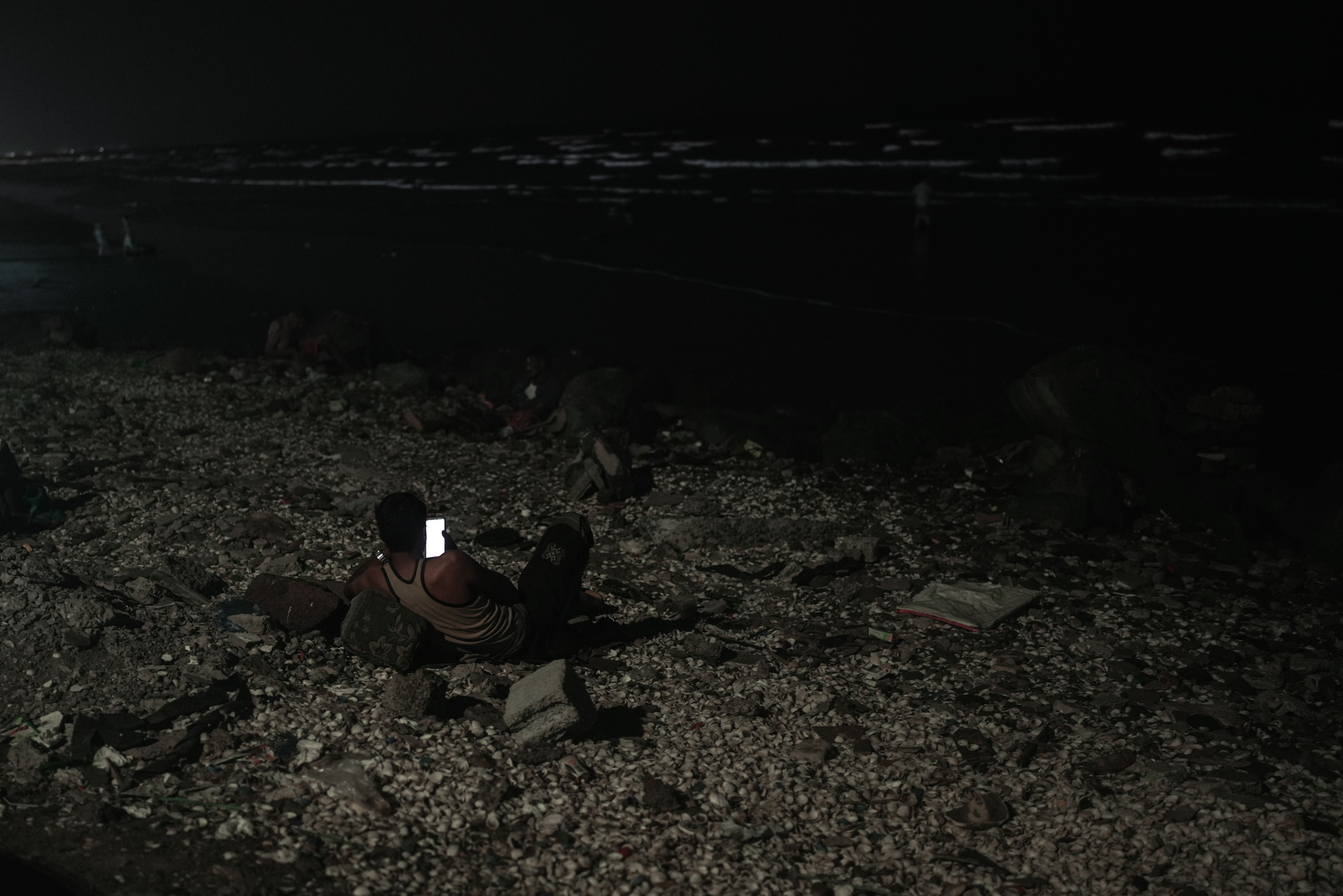 A young man rests near the sea at night, using his phone in Aden. Some residents spend hours along the coast waiting for temperatures to ease or for electricity to return to their homes. Photo: Belal Khaled